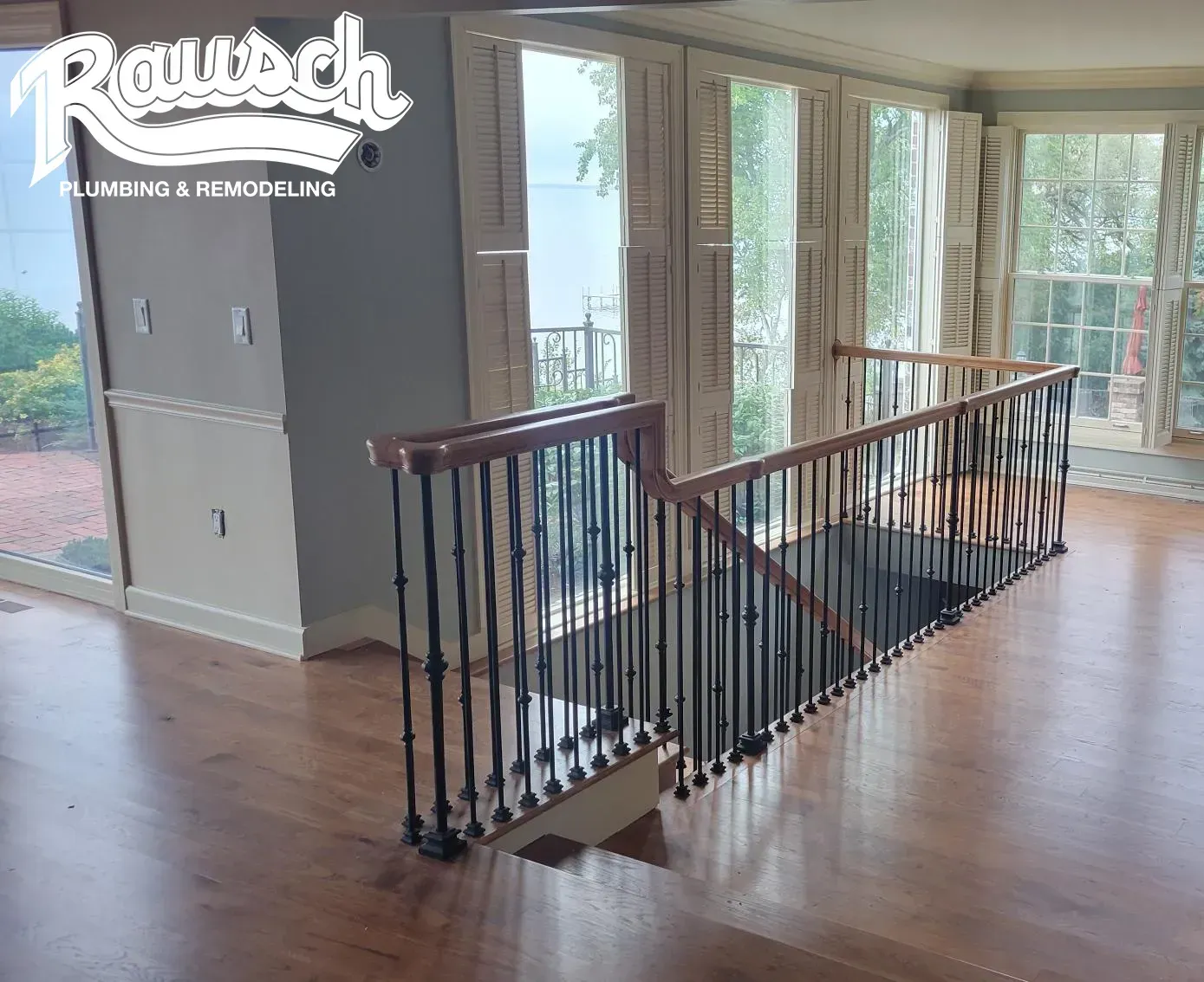 Staircase with wooden handrails and black iron spindles, leading to a room with large windows and hardwood floors.