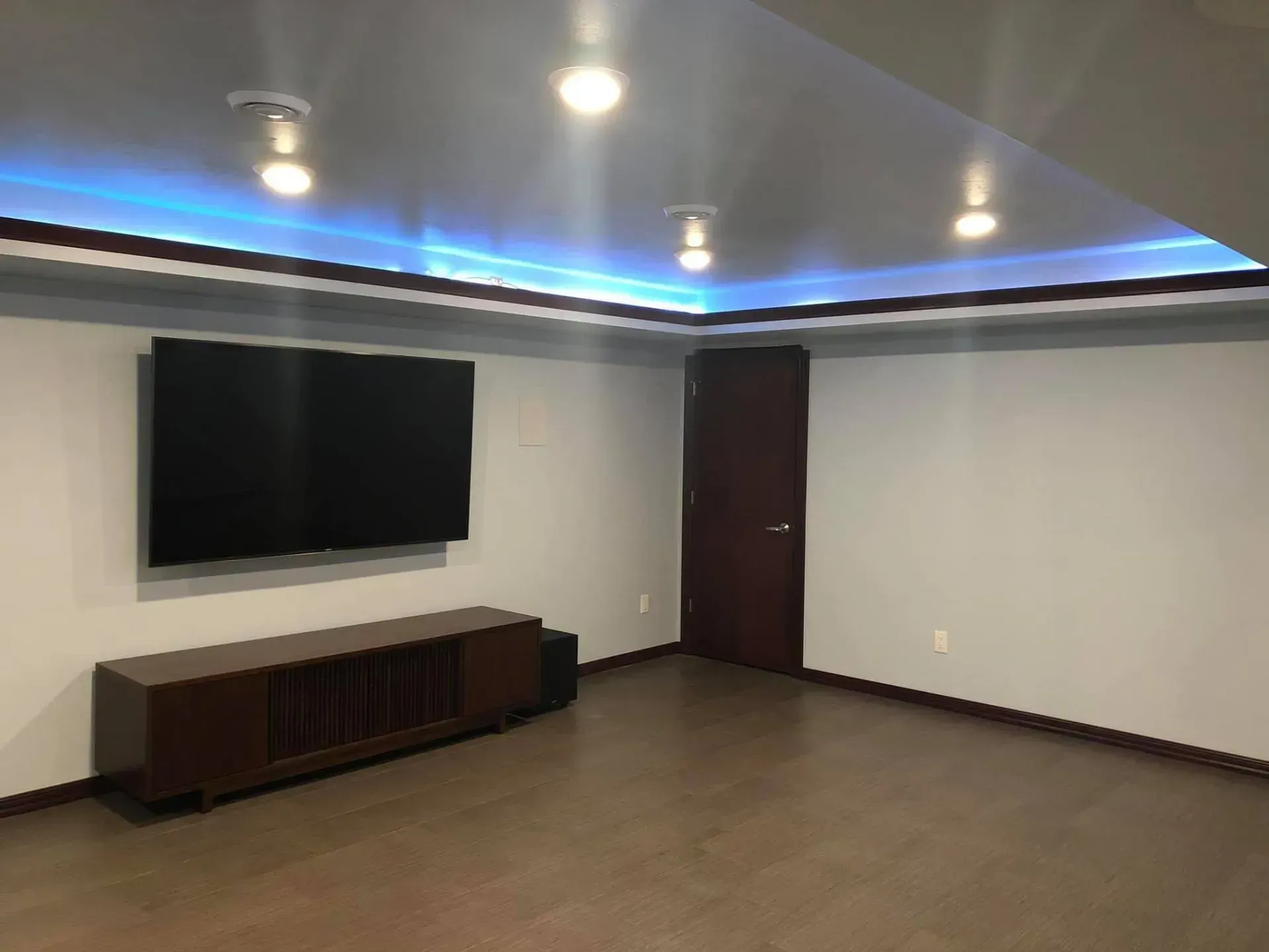 A modern basement room with a wall-mounted TV, wooden entertainment unit, and a door. Blue LED lights line the ceiling.