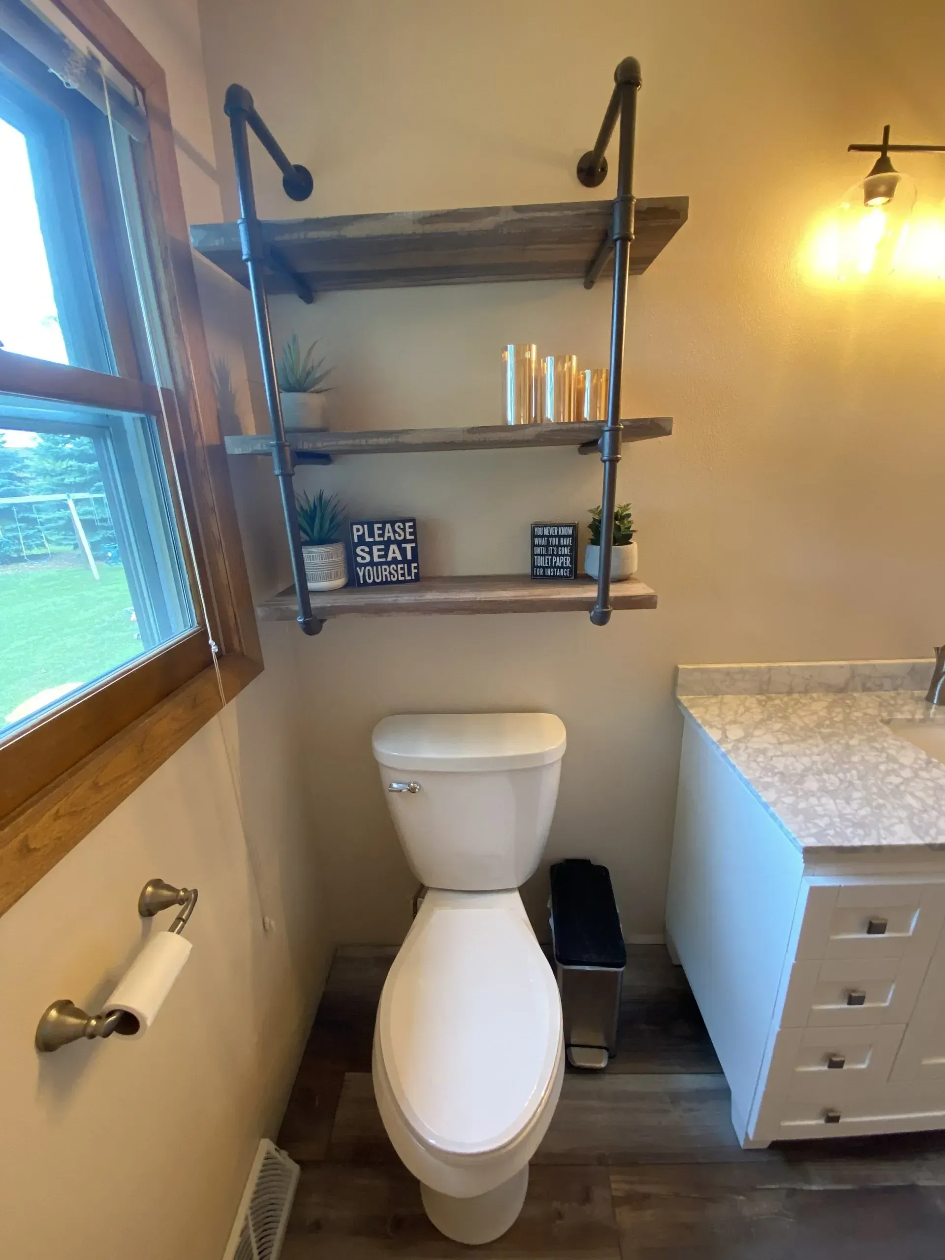 Bathroom with toilet under wood shelves, near window and vanity.