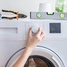 Hand turning dial on a washing machine, tools and level on shelf above.