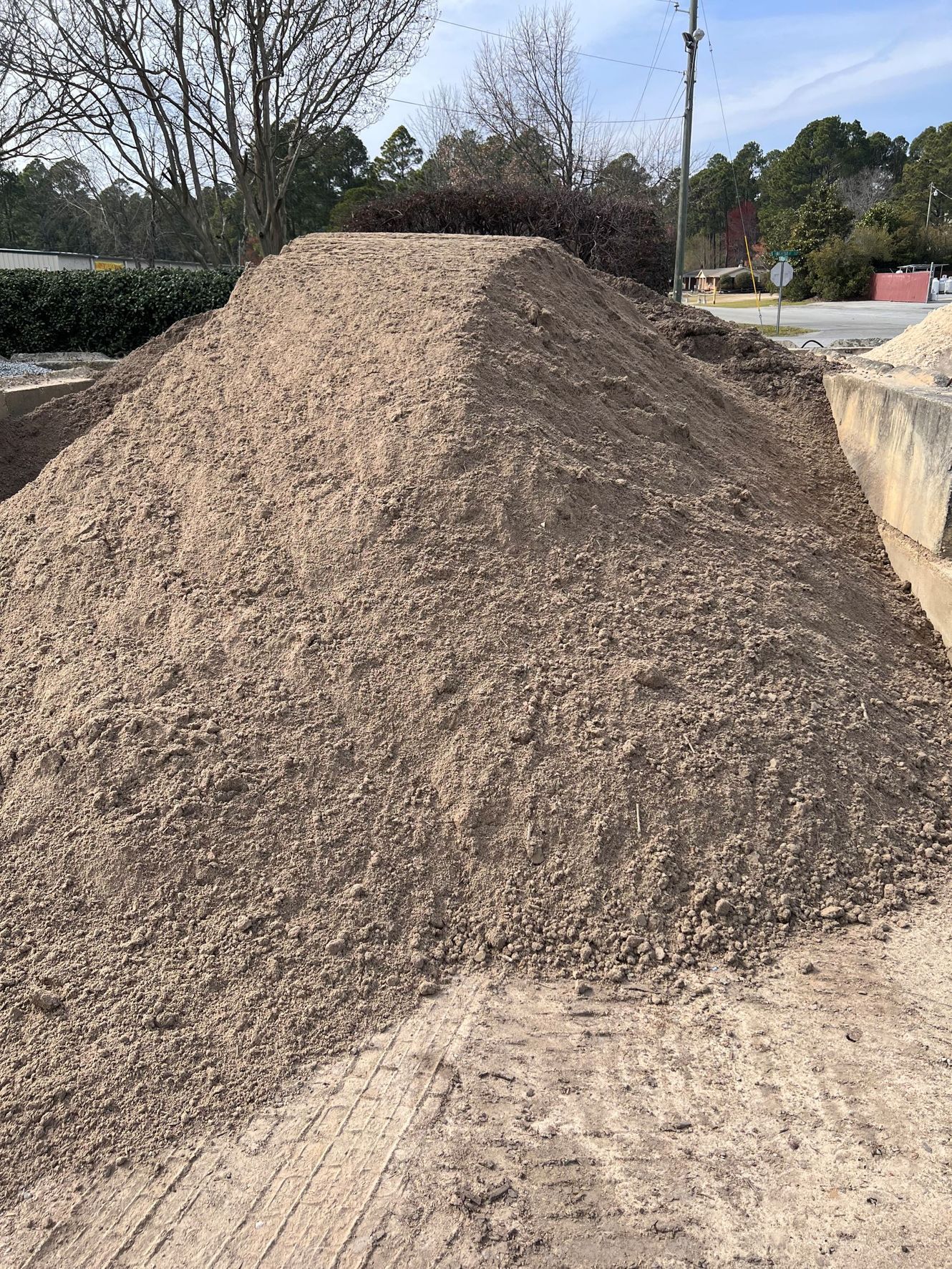 A large, conical pile of coarse, light-brown sandy soil sits in an outdoor area near trees and a paved surface.