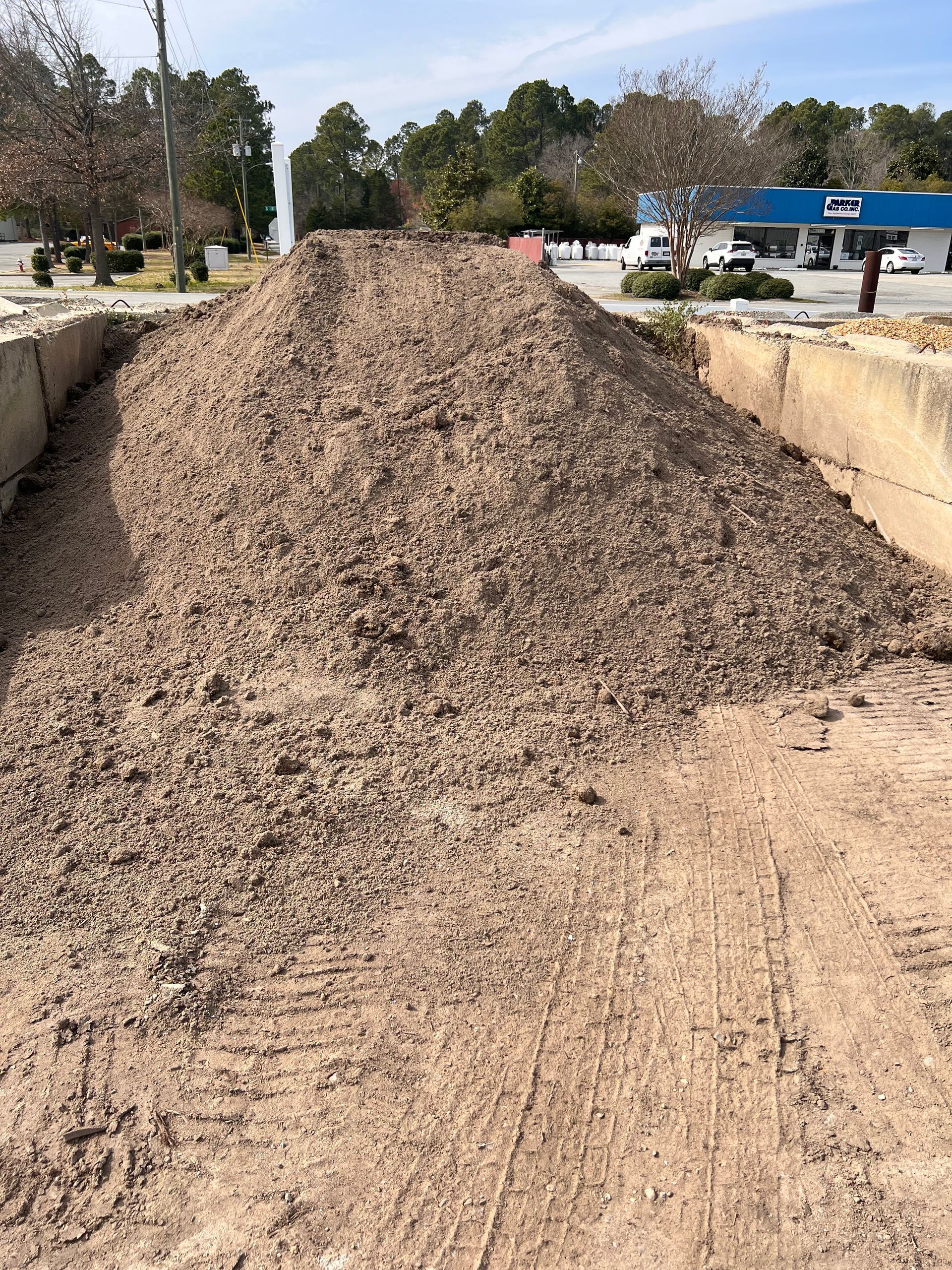 A large, conical pile of brown, coarse mulch sits between two concrete barriers outdoors on a sunny day.