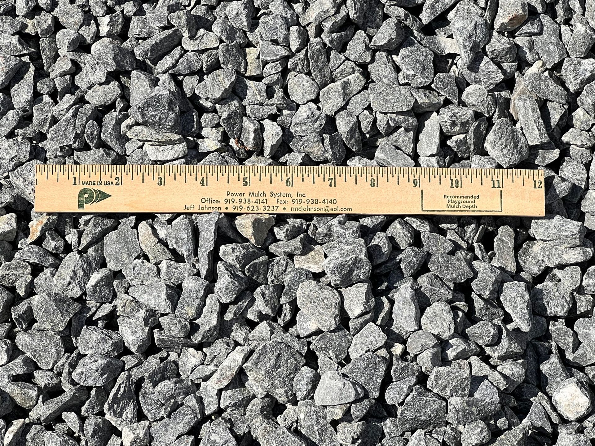 A wooden ruler lies across a bed of grey crushed stone, measuring the size of the aggregate.