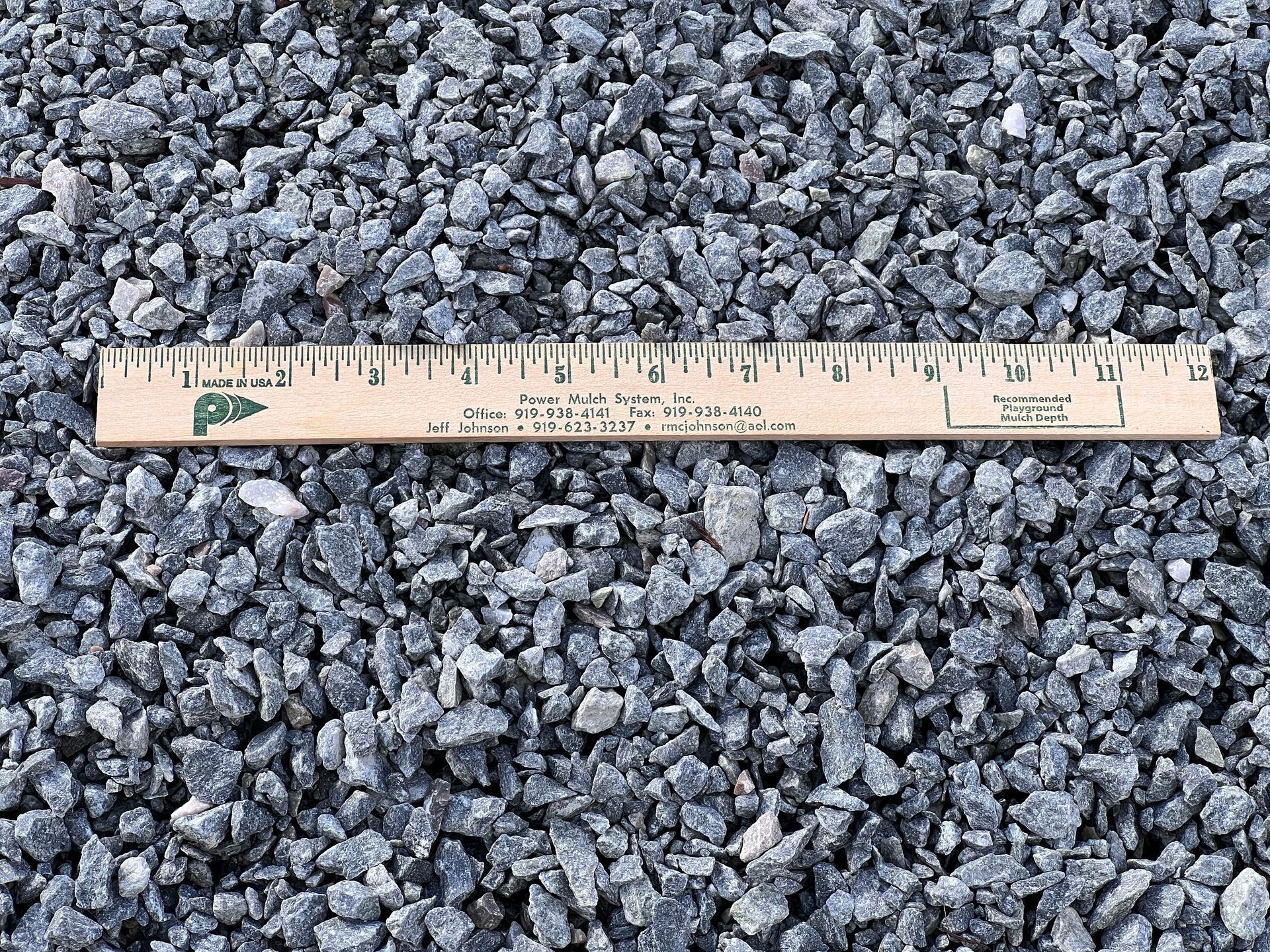 A wooden ruler rests horizontally atop a pile of gray, crushed stone aggregate.