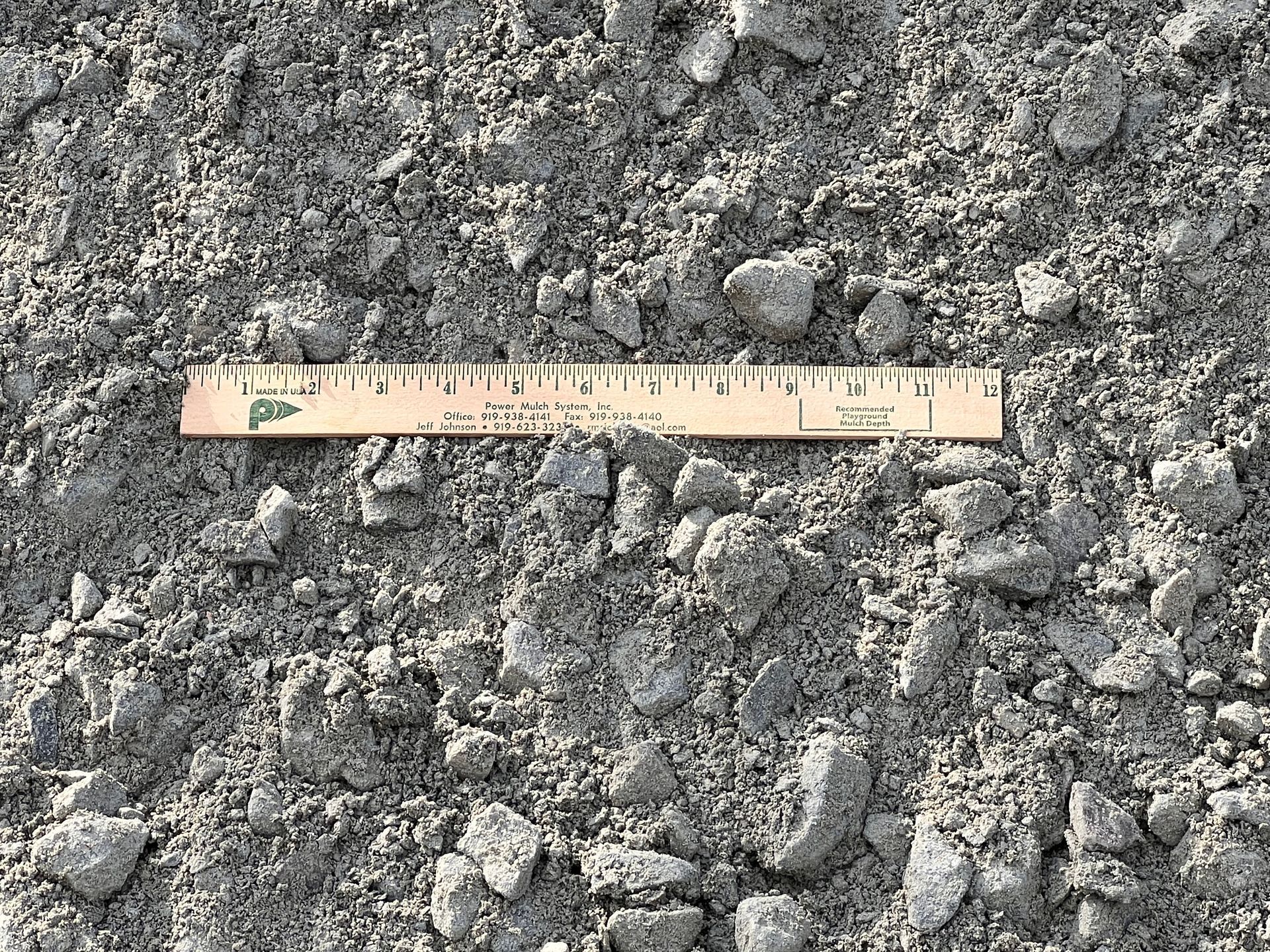 A 6-inch wooden ruler resting on a bed of loose, grey crushed gravel and dirt.