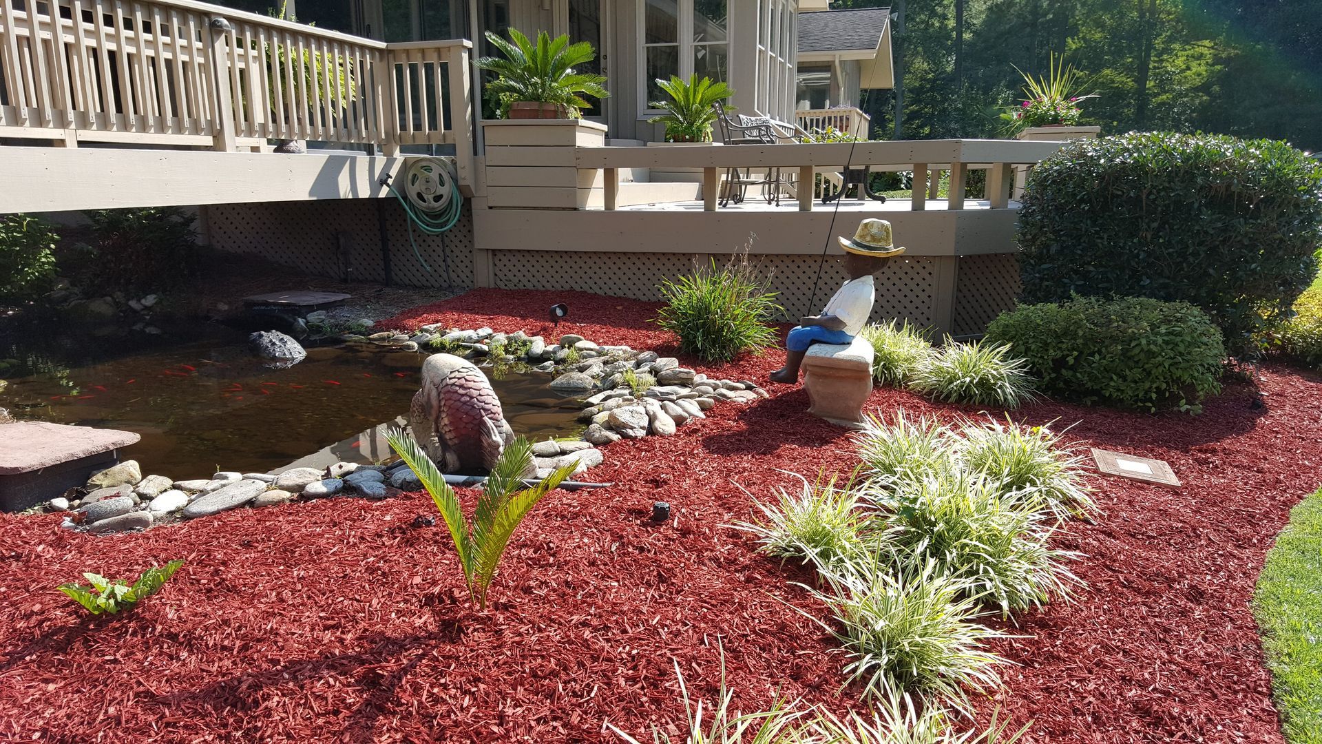 Landscaped yard with red mulch, pond, and deck of a house. A statue sits on the pond's edge.