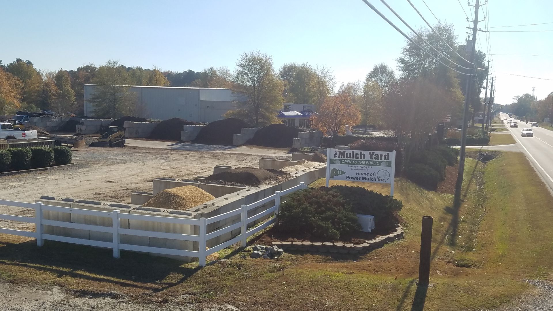 Landscape supply business with white fence, sign, and materials, next to a road under a sunny sky.