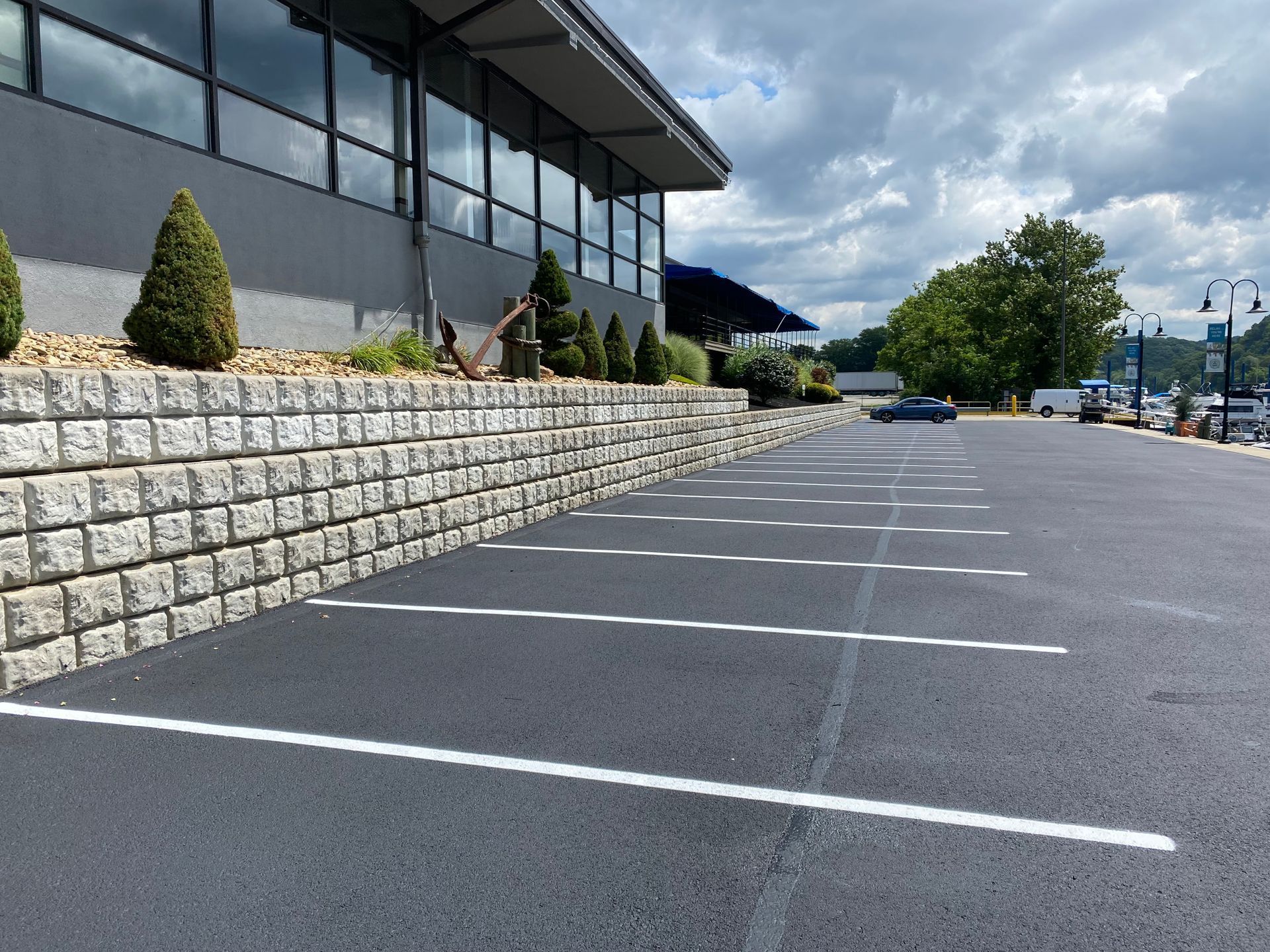 An asphalt parking lot with white-painted lines beside a tiered stone retaining wall and a modern office building.