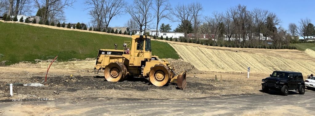 A yellow construction roller sits on a dirt lot with a grassy hill in the background, near a parked black SUV.