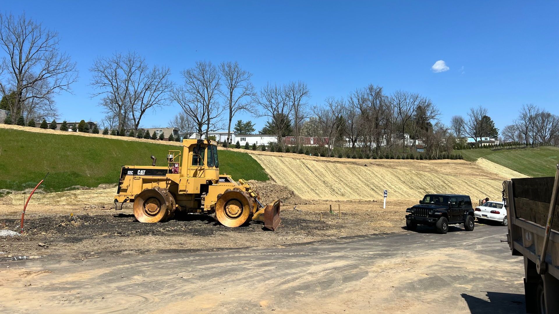 A yellow CAT construction compactor sits on a dirt lot near a sloped, straw-covered embankment under a clear blue sky.