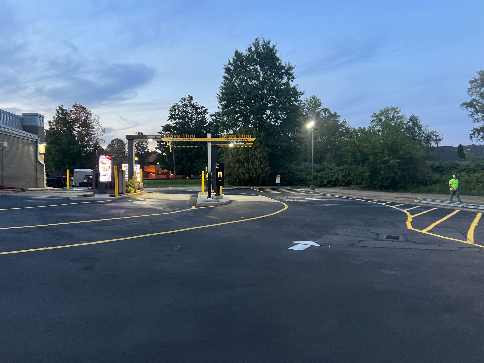 Drive-thru lane with a yellow height clearance bar and menu boards in a parking lot during twilight.
