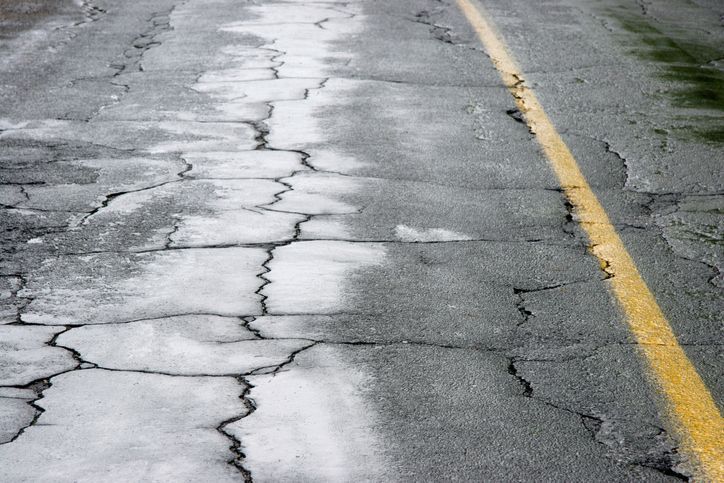 A weathered asphalt road surface with extensive cracking and a yellow painted shoulder line.