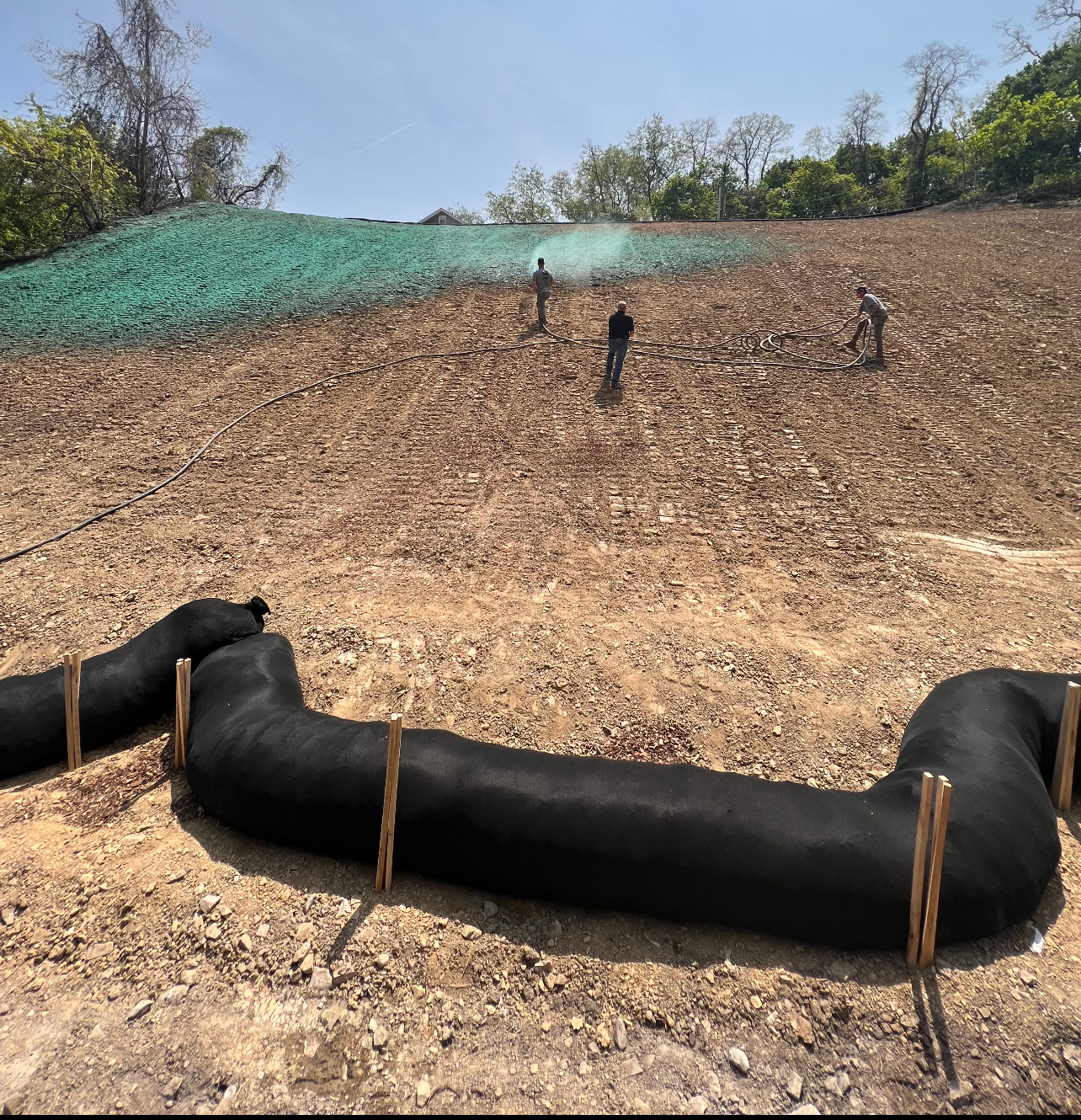 Workers perform hydroseeding on a hillside slope protected by black erosion control wattles secured with wooden stakes.
