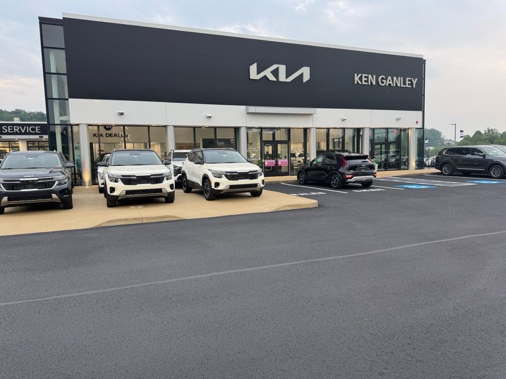 The Ken Ganley Kia dealership building with several cars parked out front on a paved lot under a cloudy sky.