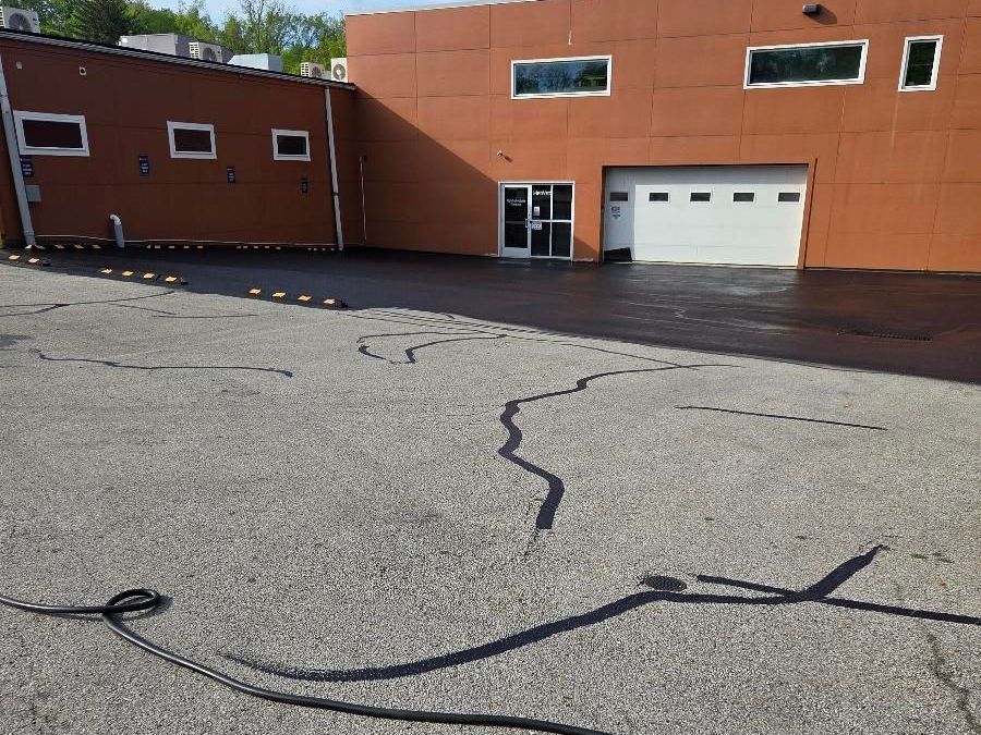 A tan industrial building with a white garage door, adjacent to an asphalt parking lot with black crack-seal lines.