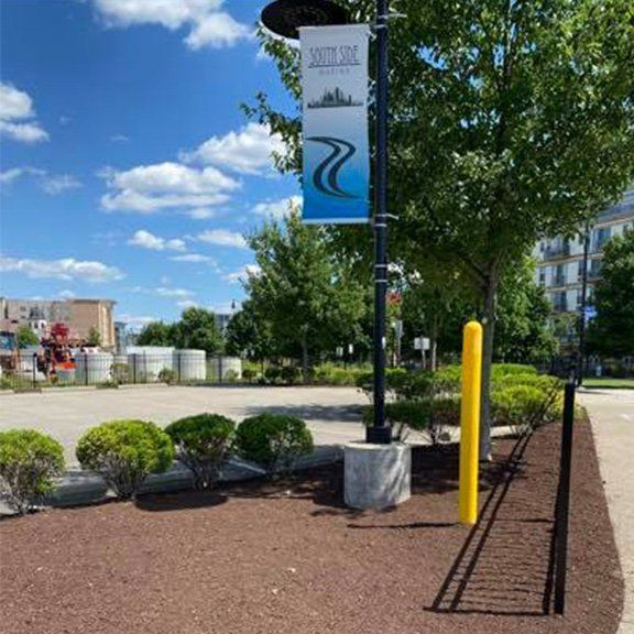 A banner displaying a blue river logo hangs from a pole near green bushes, mulch, and a yellow safety post under a blue sky.