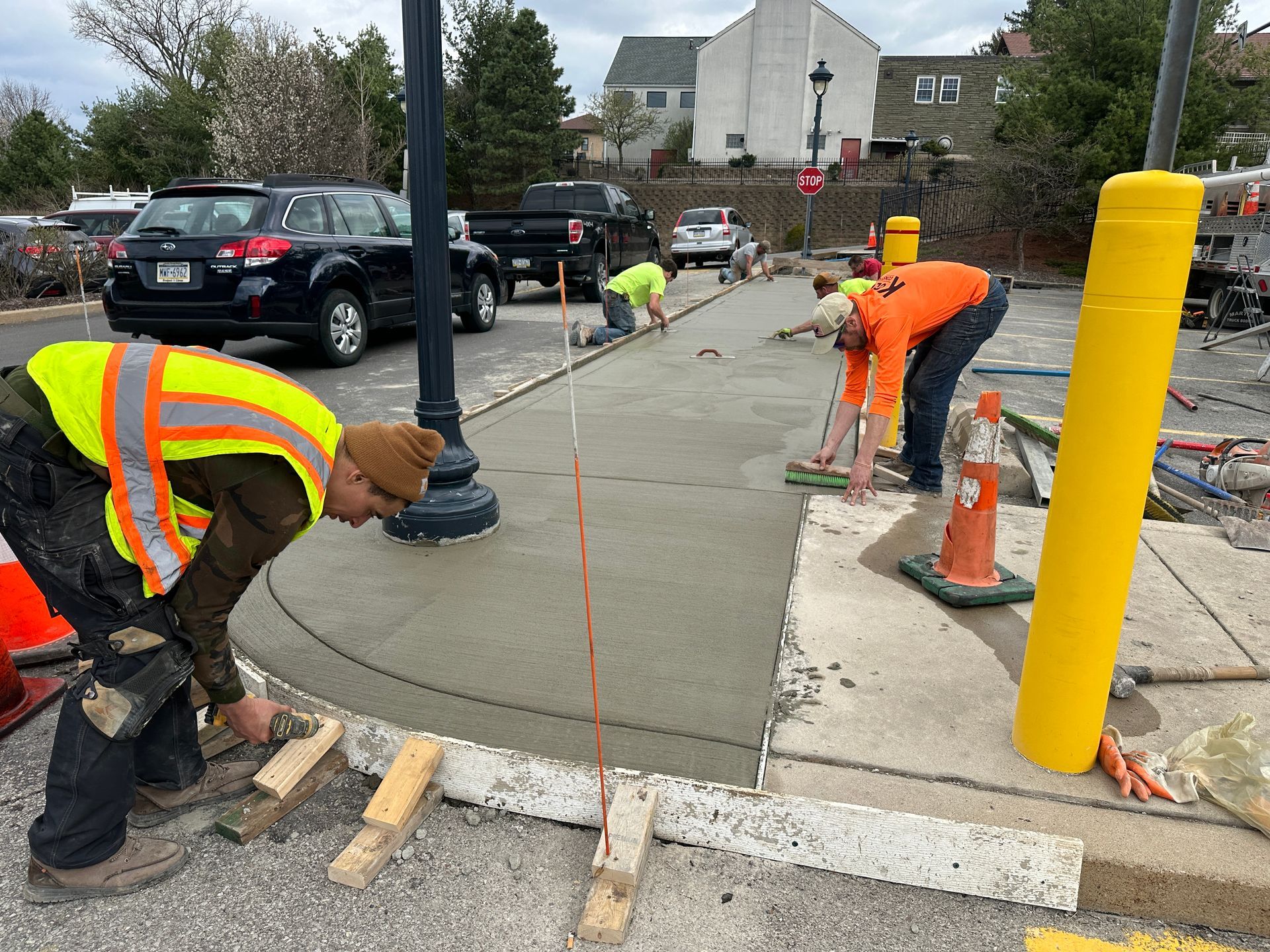 Road workers pouring concrete and smoothing a sidewalk beside orange cones and a yellow bollard