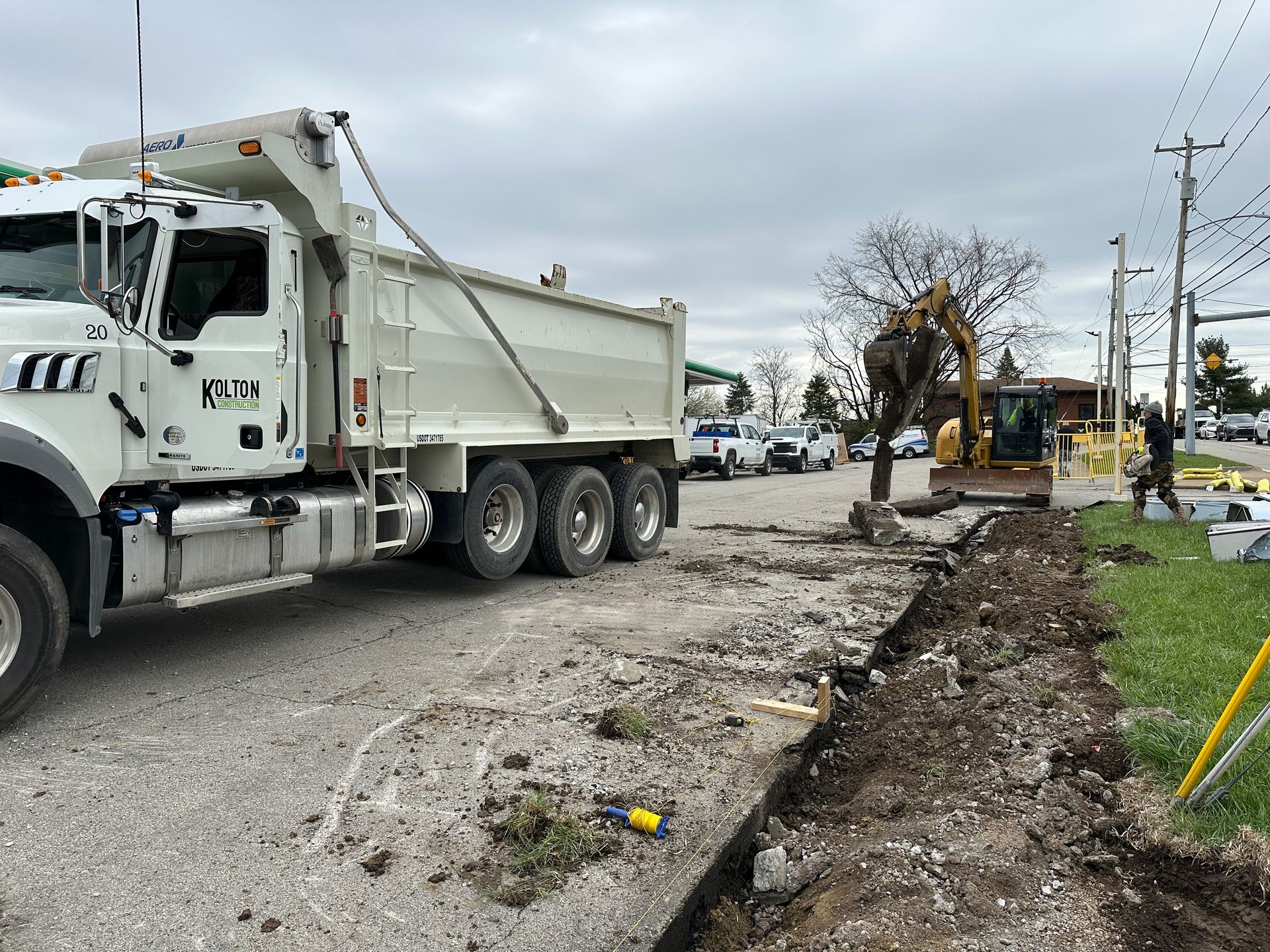 White dump truck beside a torn-up roadside trench with construction equipment and workers in the distance