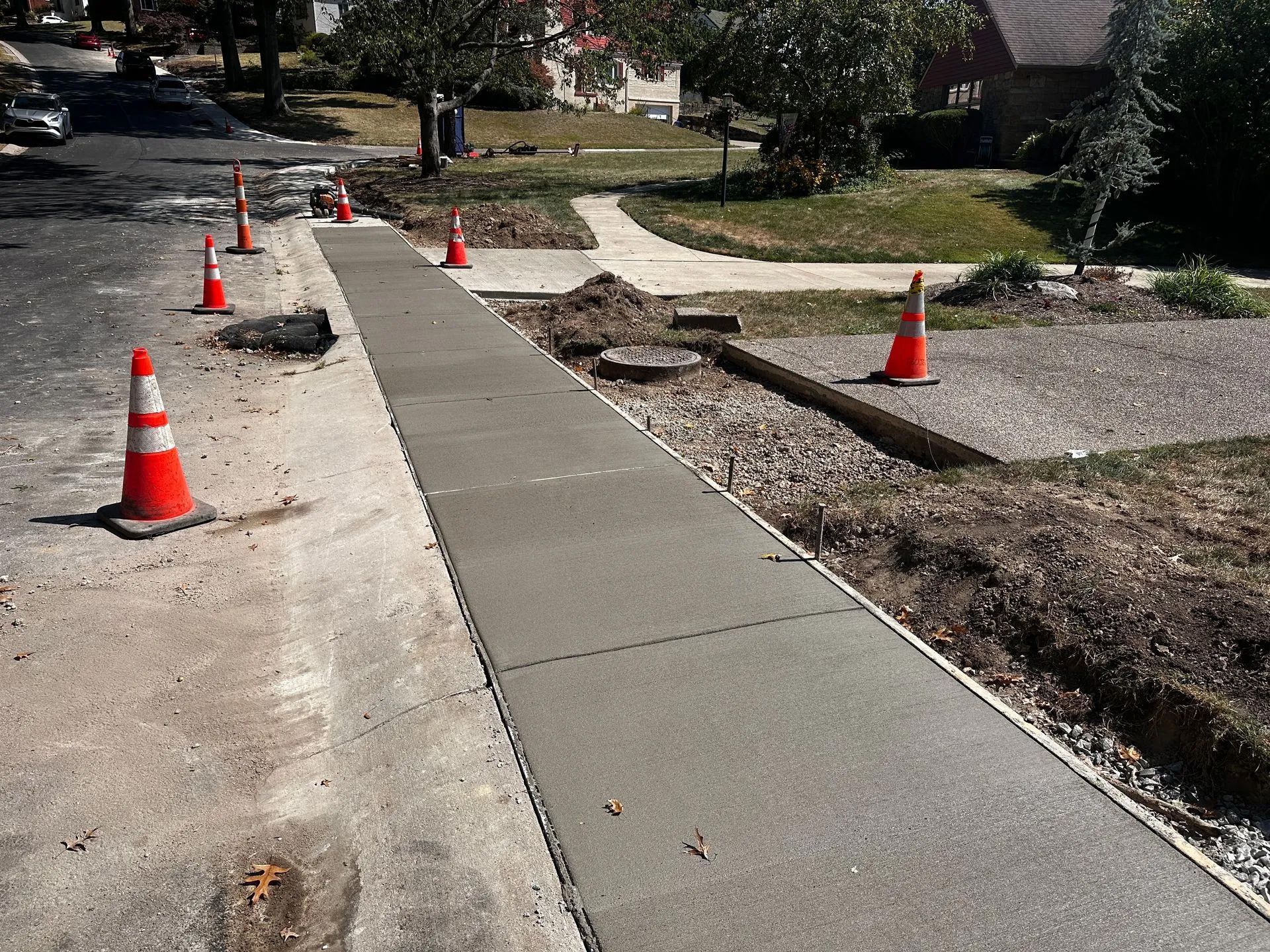 Freshly poured concrete sidewalk sections line a residential street with orange traffic safety cones.