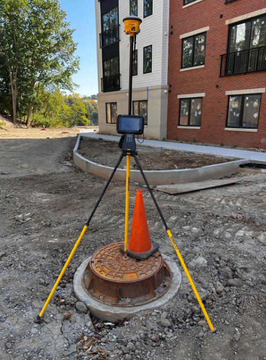 Survey camera on tripod beside orange cone at a construction site outside a brick building.