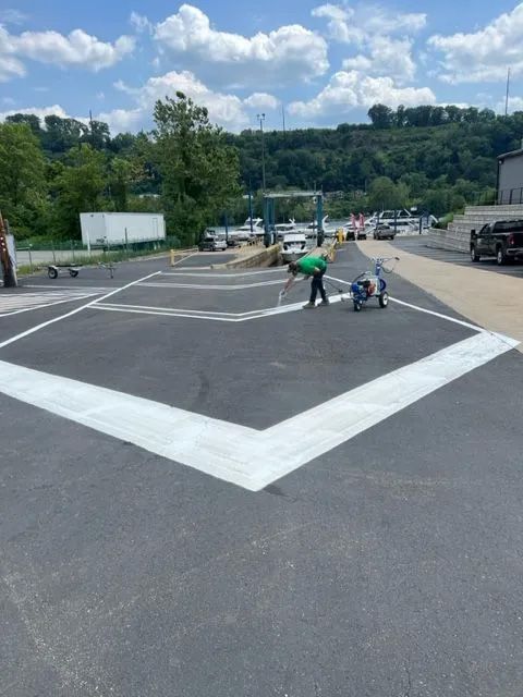 A worker paints white chevron markings on an asphalt parking lot under a bright, cloudy sky.