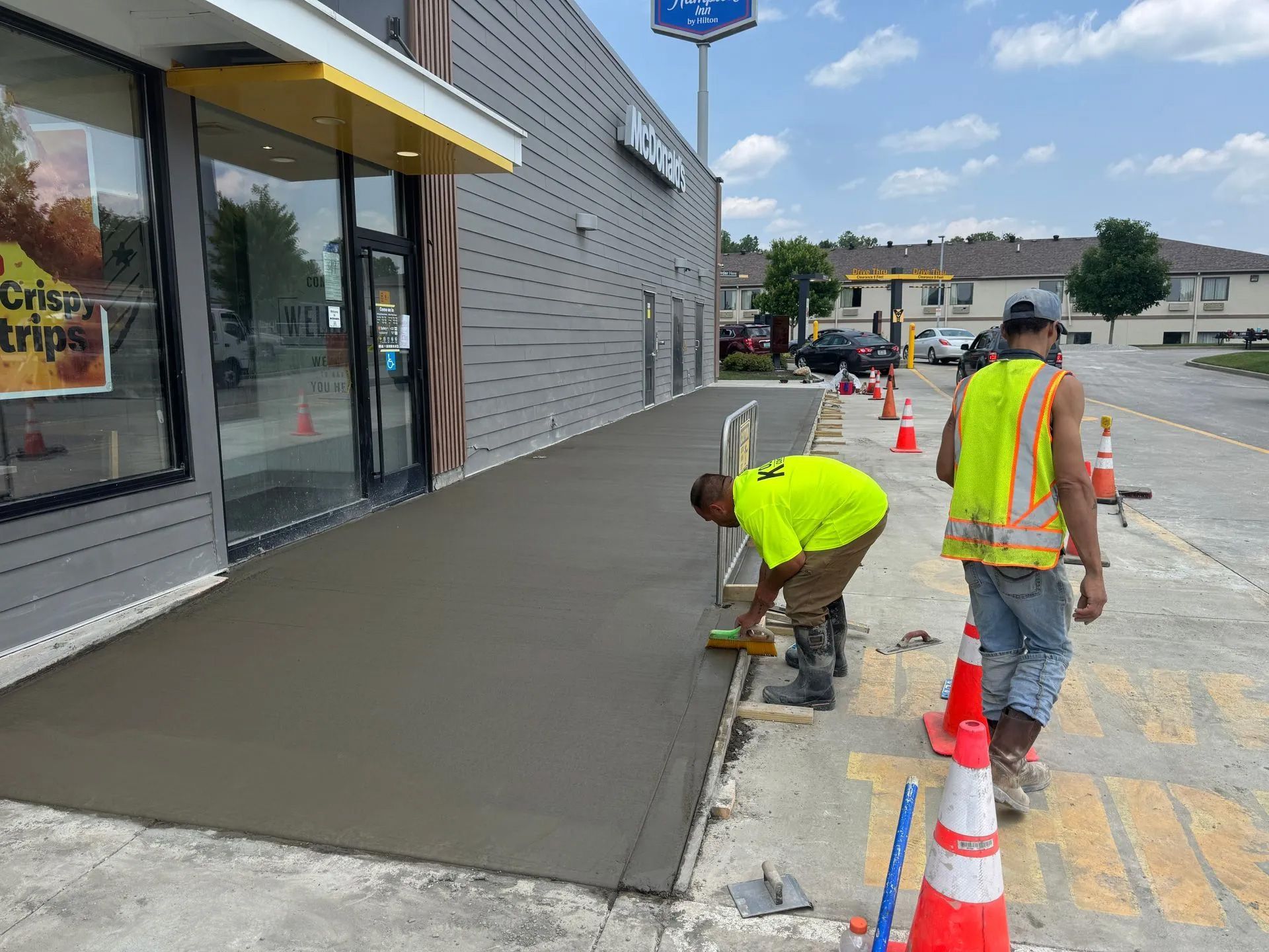 Workers pouring fresh concrete on a sidewalk beside a storefront, with cones marking the area.