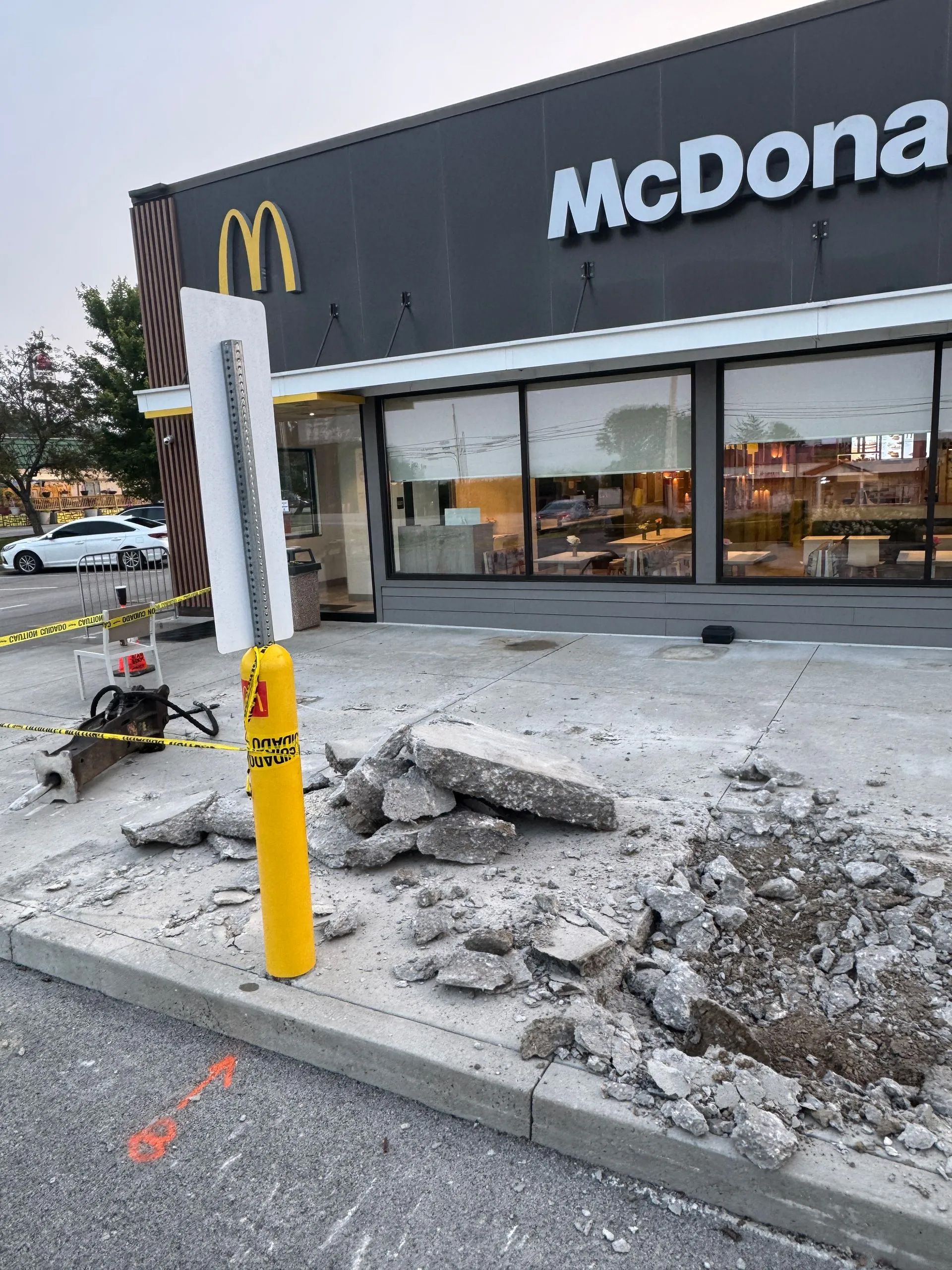 Construction work in front of a McDonald’s entrance with concrete rubble, a yellow bollard, and caution tape.