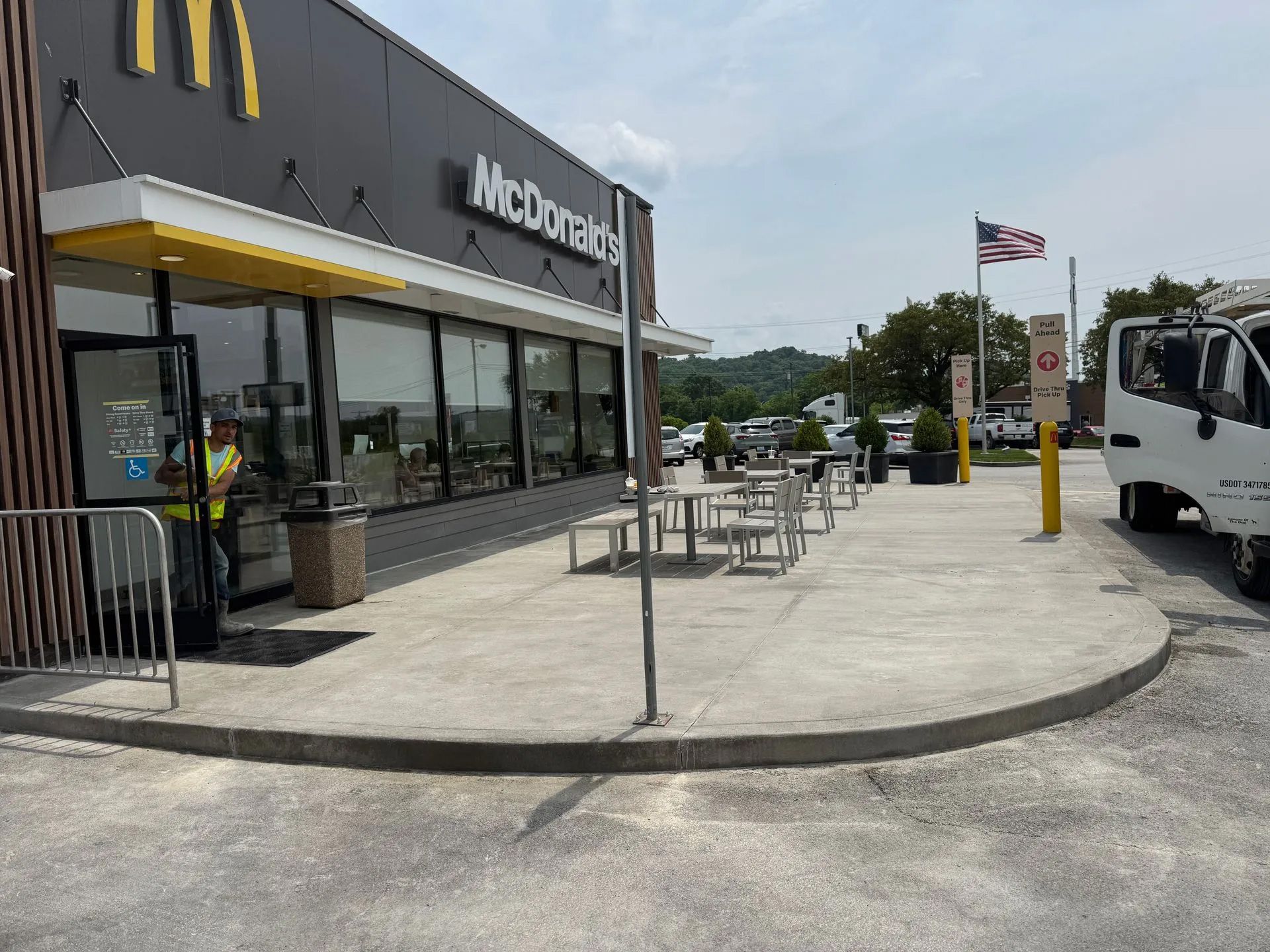 A modern McDonald's restaurant exterior with outdoor seating, a metal railing, and a parked white truck on a sunny day.