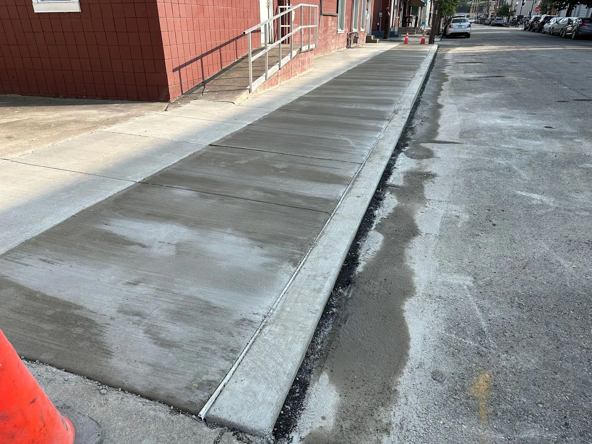 Newly poured concrete sidewalk with a metal curb edge running alongside a red brick building and an asphalt street.