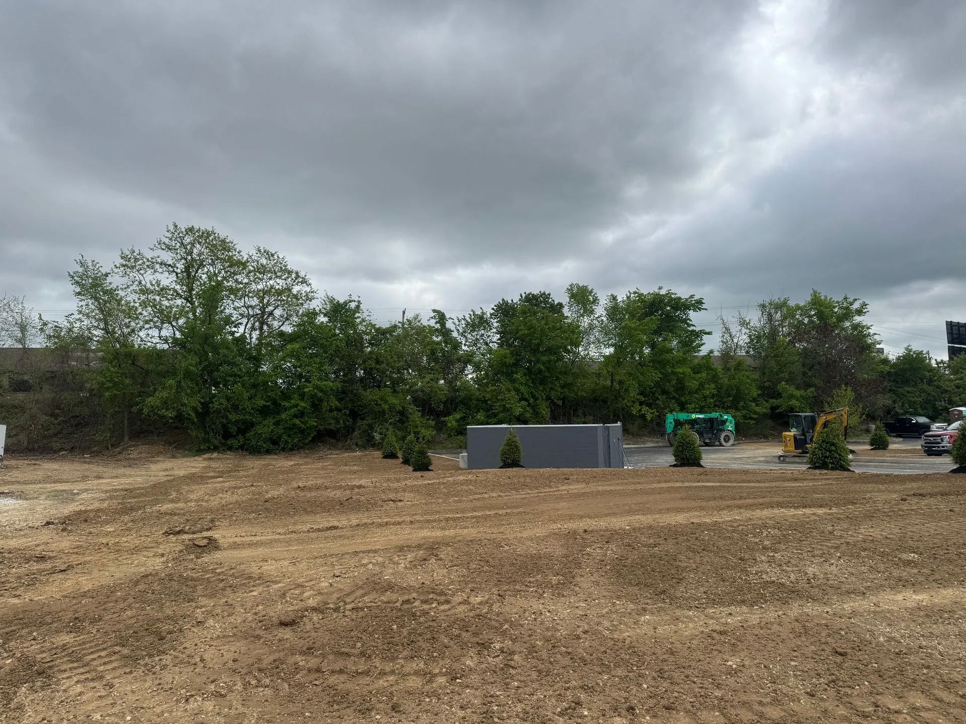 A vacant, dirt-covered construction lot with a grey utility structure and green trees under a cloudy sky.