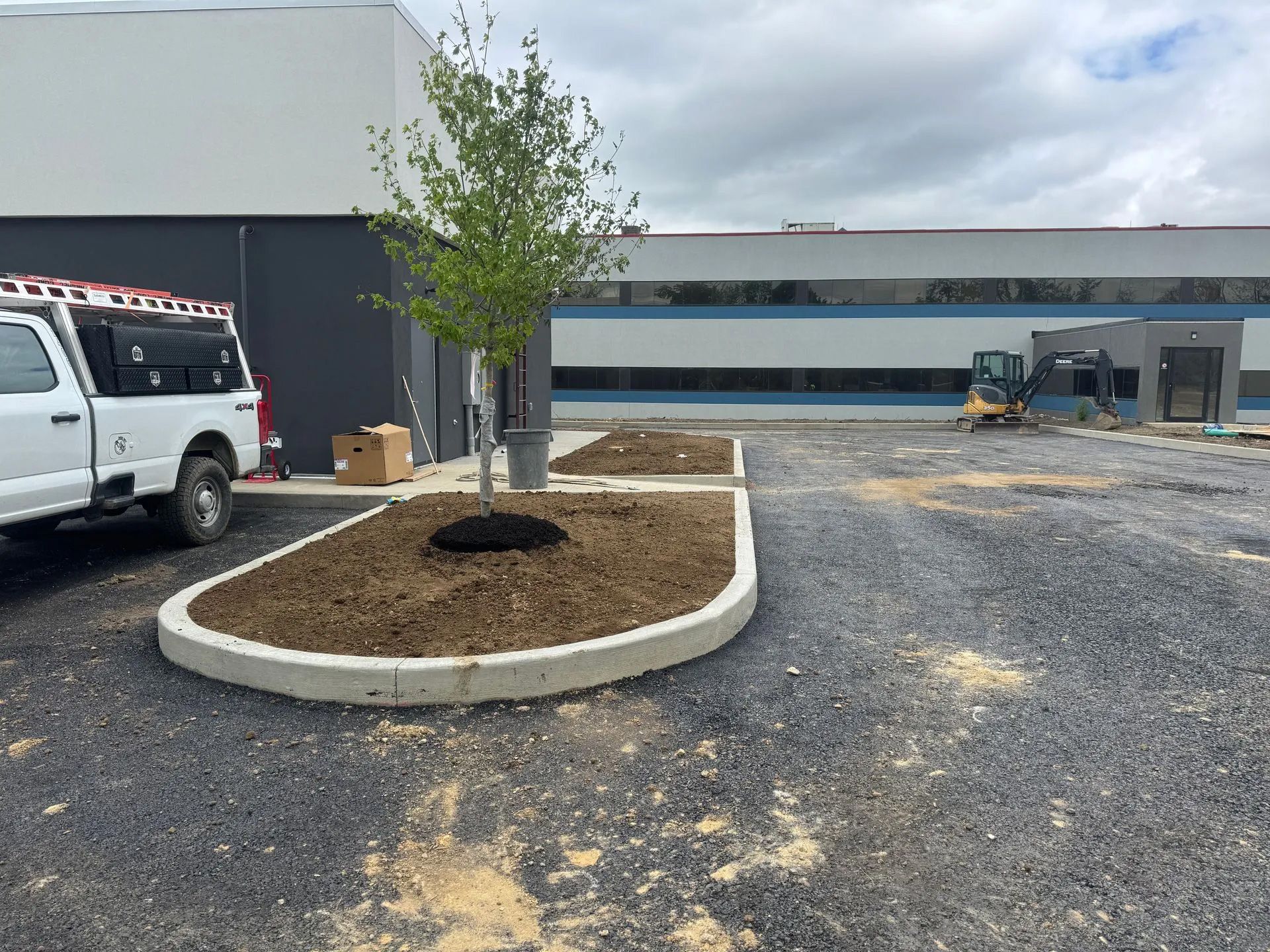 A white utility truck is parked next to a newly landscaped island with a tree in a gravel lot in front of a commercial building.