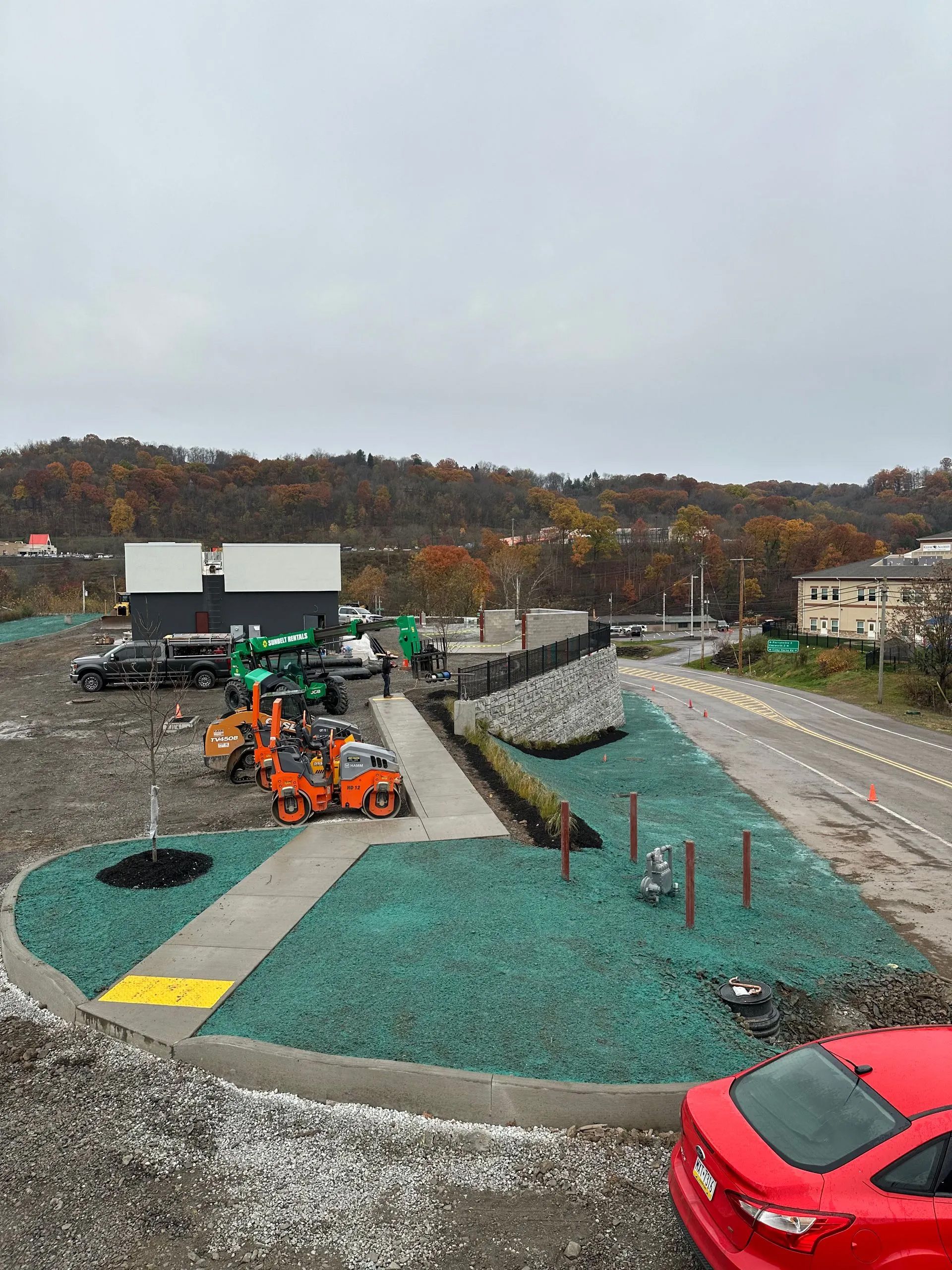Construction site with heavy machinery, a new sidewalk, and green mulch landscaping next to a road under a gray sky.
