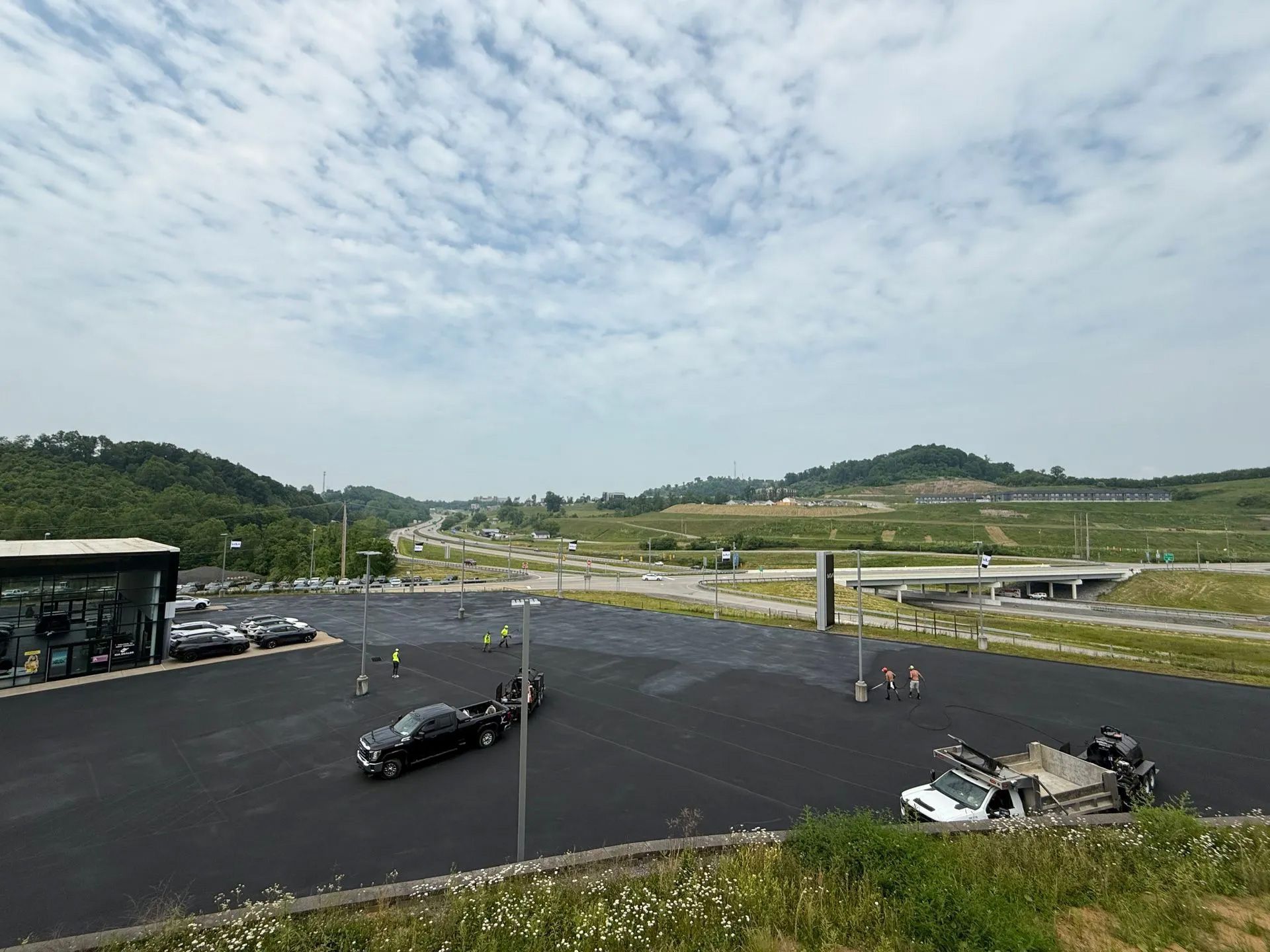 An aerial view of a paved parking lot with a few vehicles under a bright, cloudy sky with hills in the background.