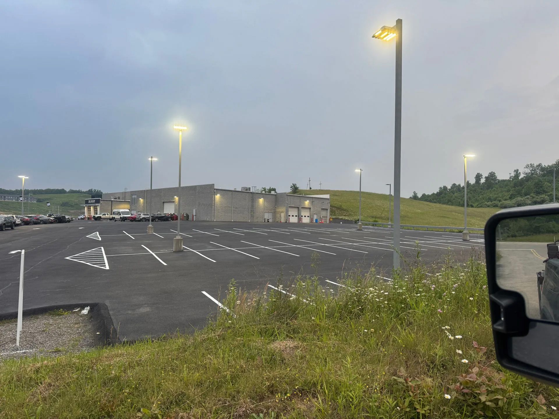 A paved parking lot with multiple light poles in front of a gray industrial building under a cloudy sky.