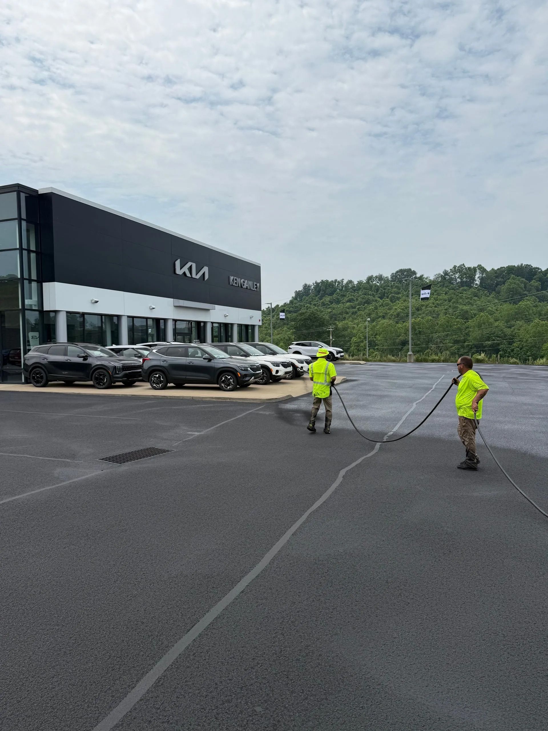 Two workers in neon vests apply sealant to a large asphalt parking lot in front of a Kia dealership.