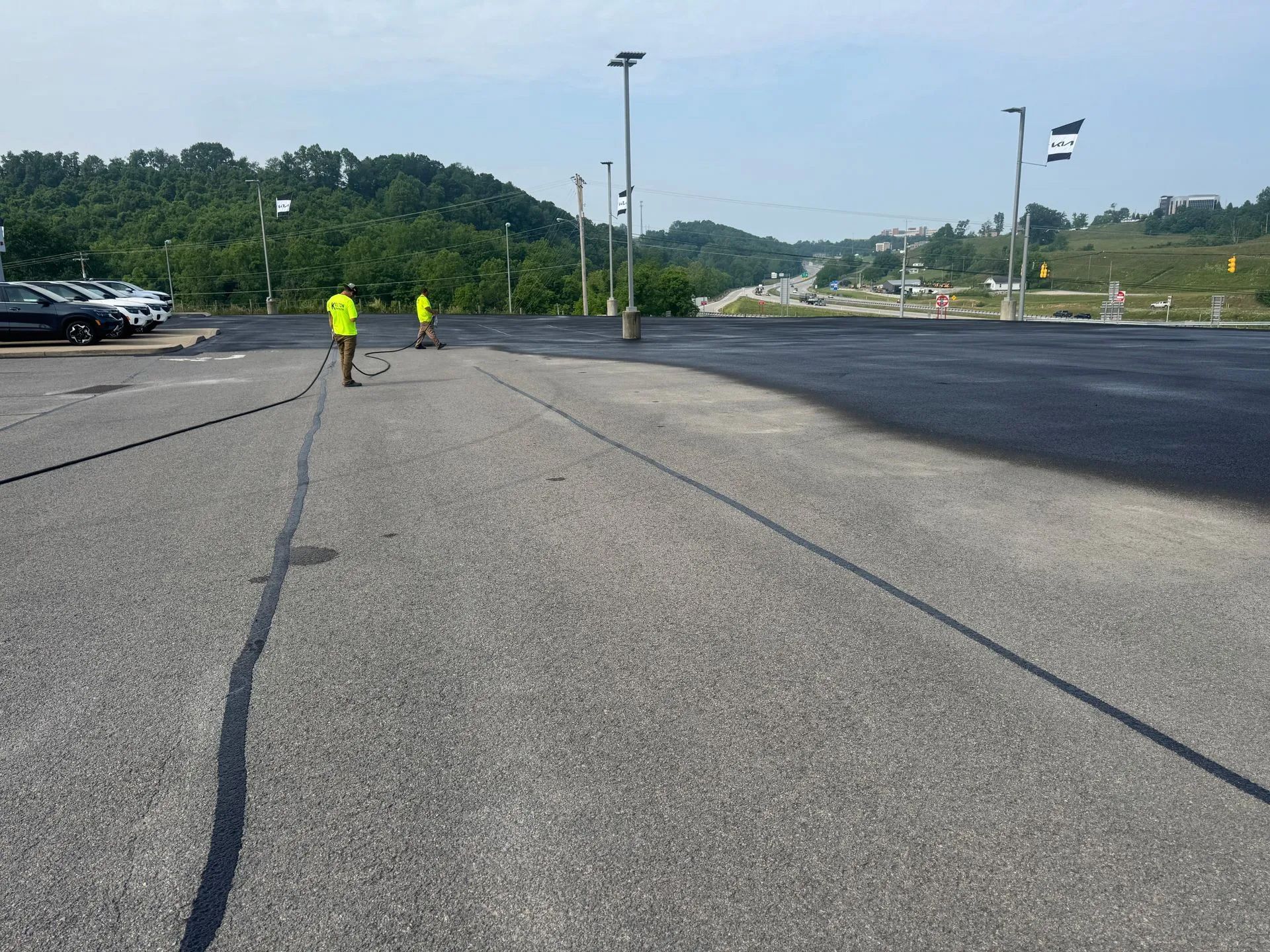Two workers in high-visibility yellow vests apply sealant to a large, paved parking lot.