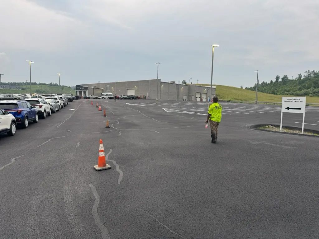 A person in a high-visibility vest walks across a large, empty asphalt parking lot toward a low concrete building.
