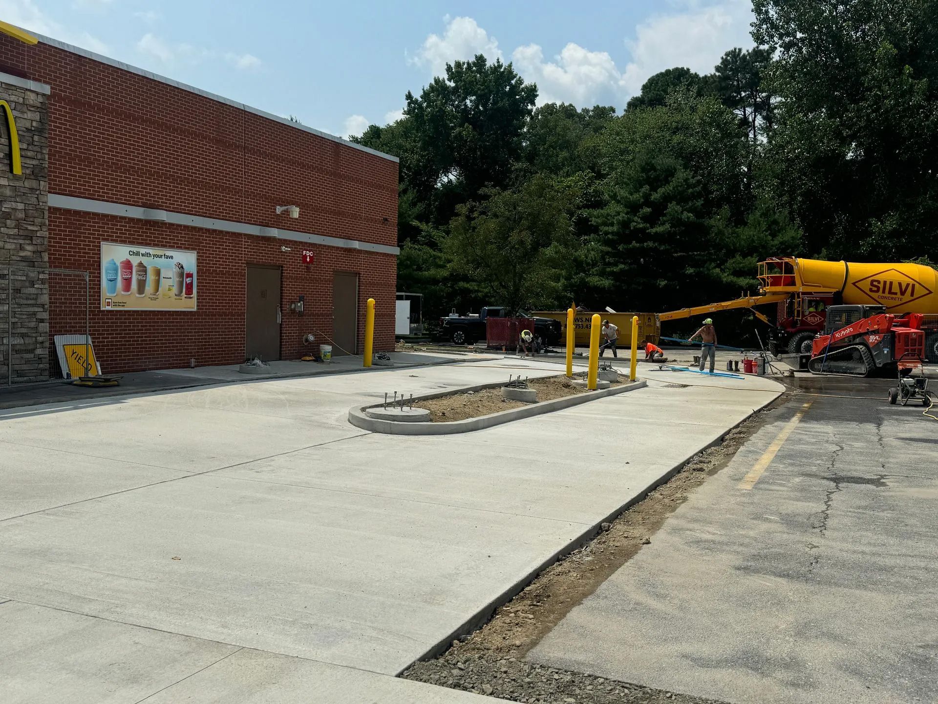 Construction workers operate a yellow cement truck outside a brick building with a concrete parking area.