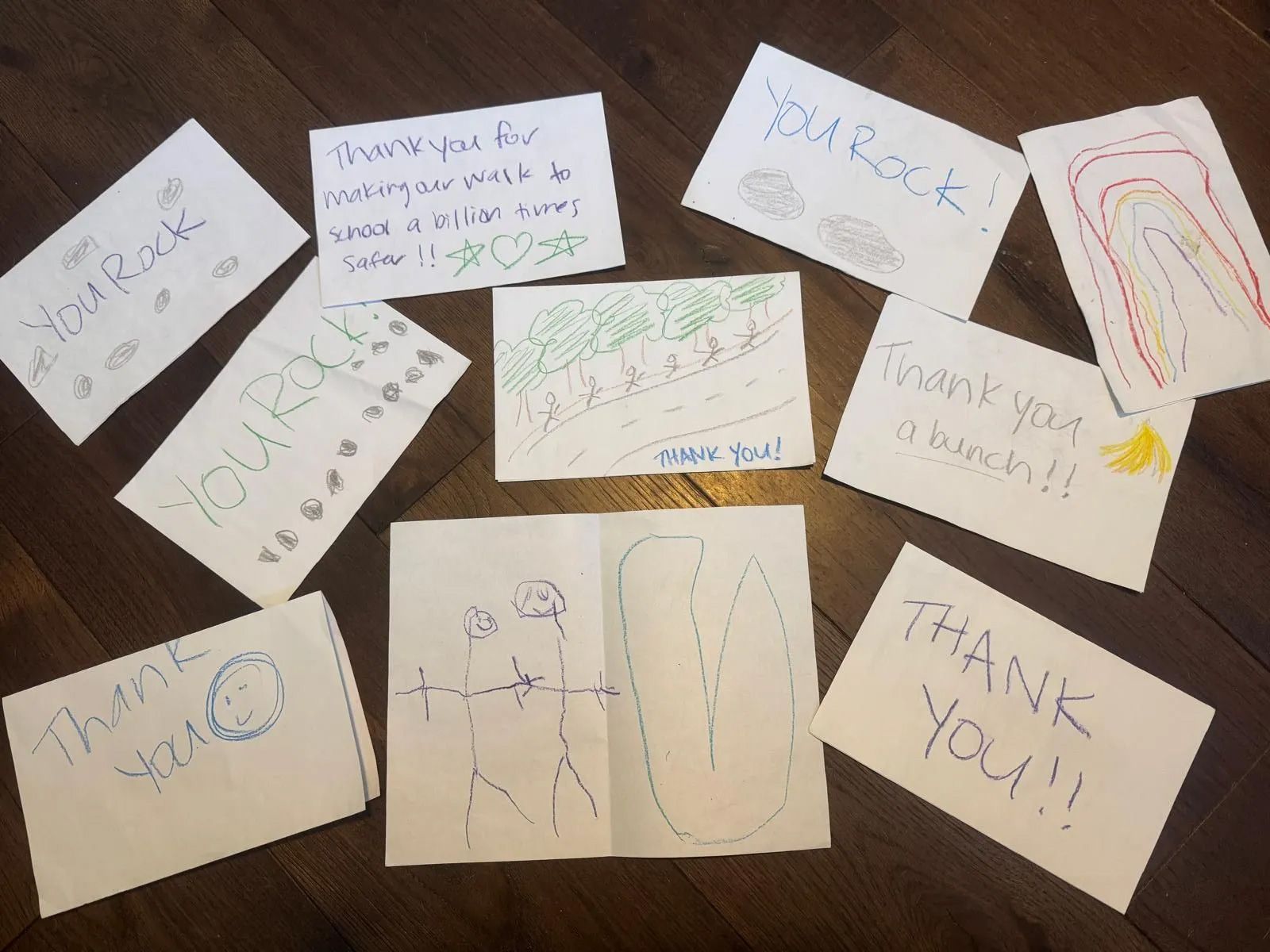 Handwritten thank-you notes and drawings decorated with stars, rainbows, and smiley faces, arranged on a wood floor.