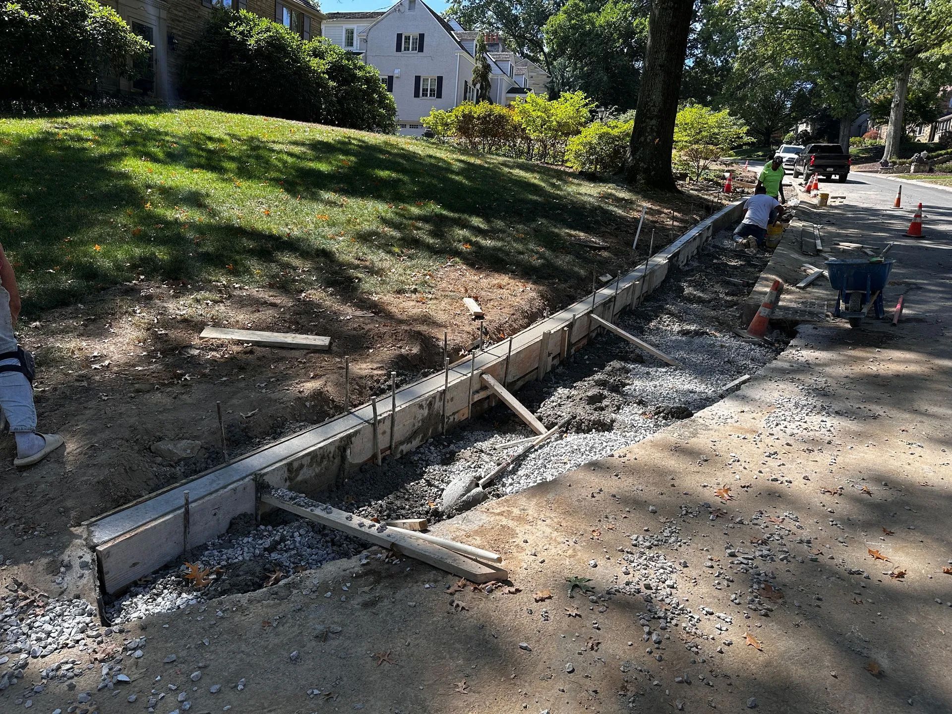 Construction workers install concrete blocks for a retaining wall along a sloped, grassy roadside.