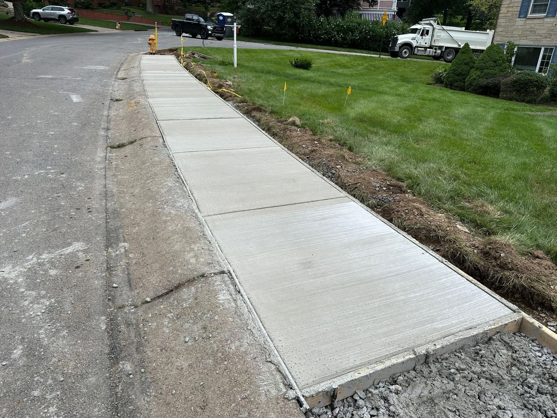A freshly poured concrete sidewalk section under construction, bordered by an asphalt road and a green grass lawn.