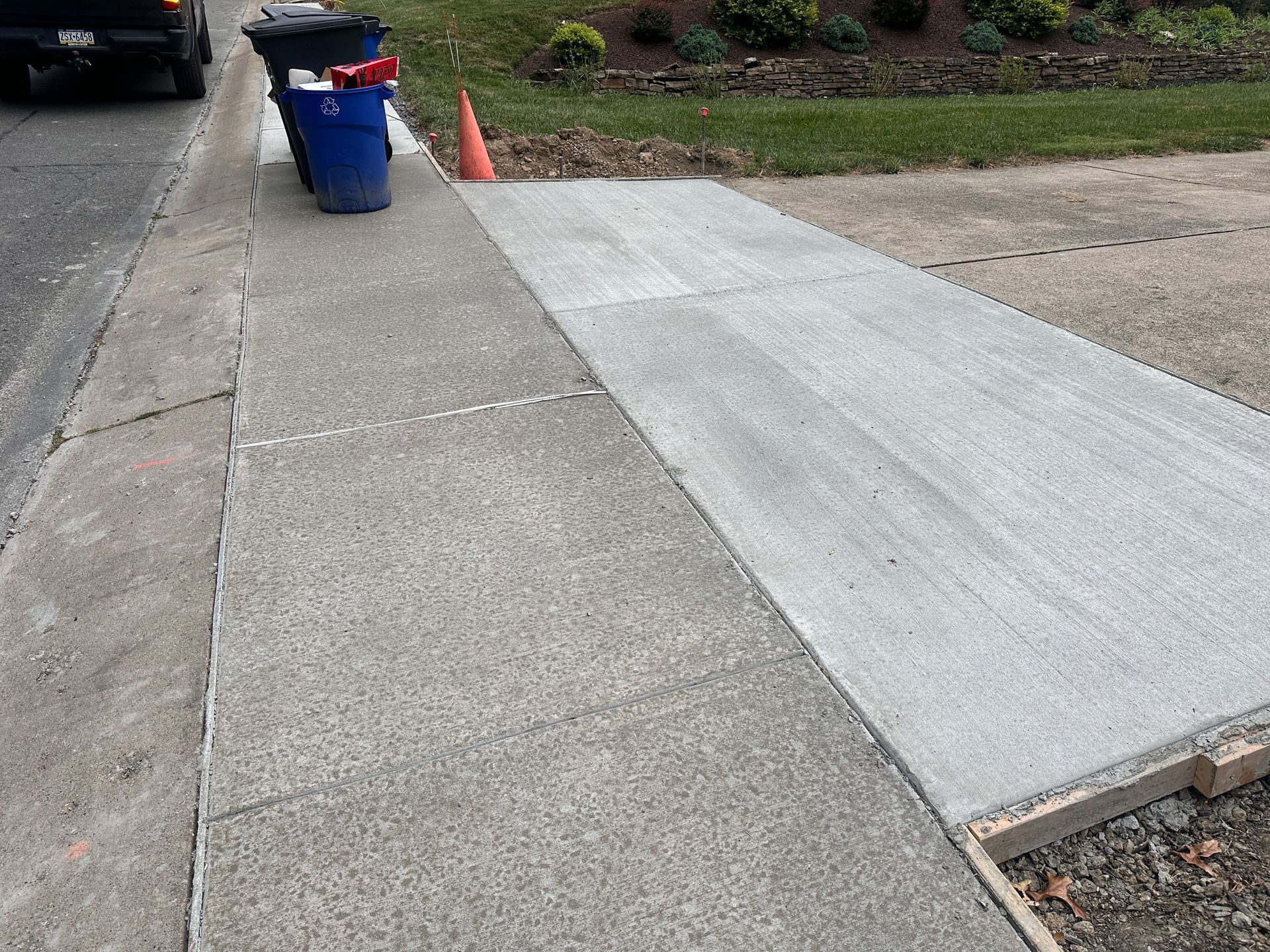 A newly poured section of concrete sidewalk sits beside an older segment, with a traffic cone and waste bins nearby.