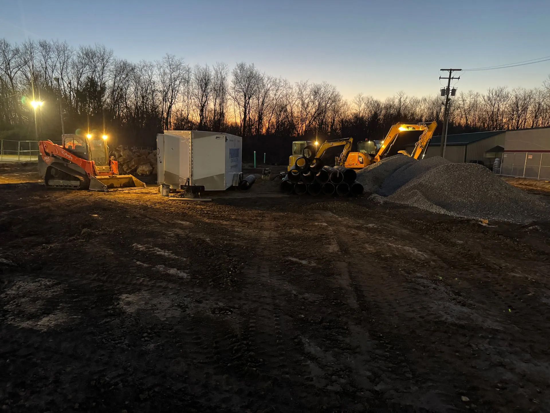 Construction site at dusk featuring heavy machinery, a portable office trailer, and a pile of gravel.
