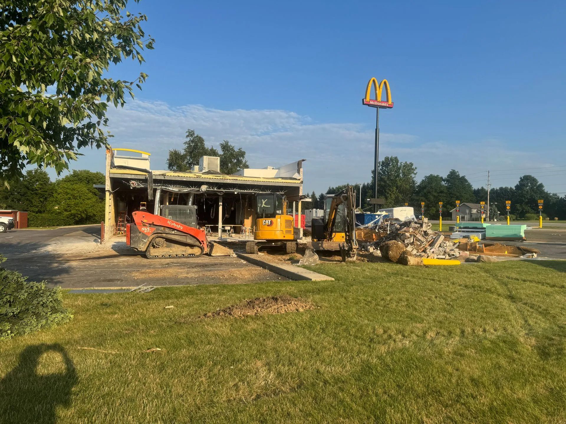 A red skid steer and a yellow excavator demolish a McDonald’s building under a clear blue sky.