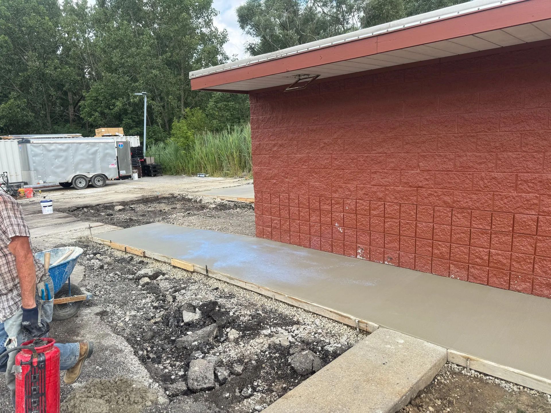 Fresh concrete walkway beside a red brick building, with construction debris and a worker at left