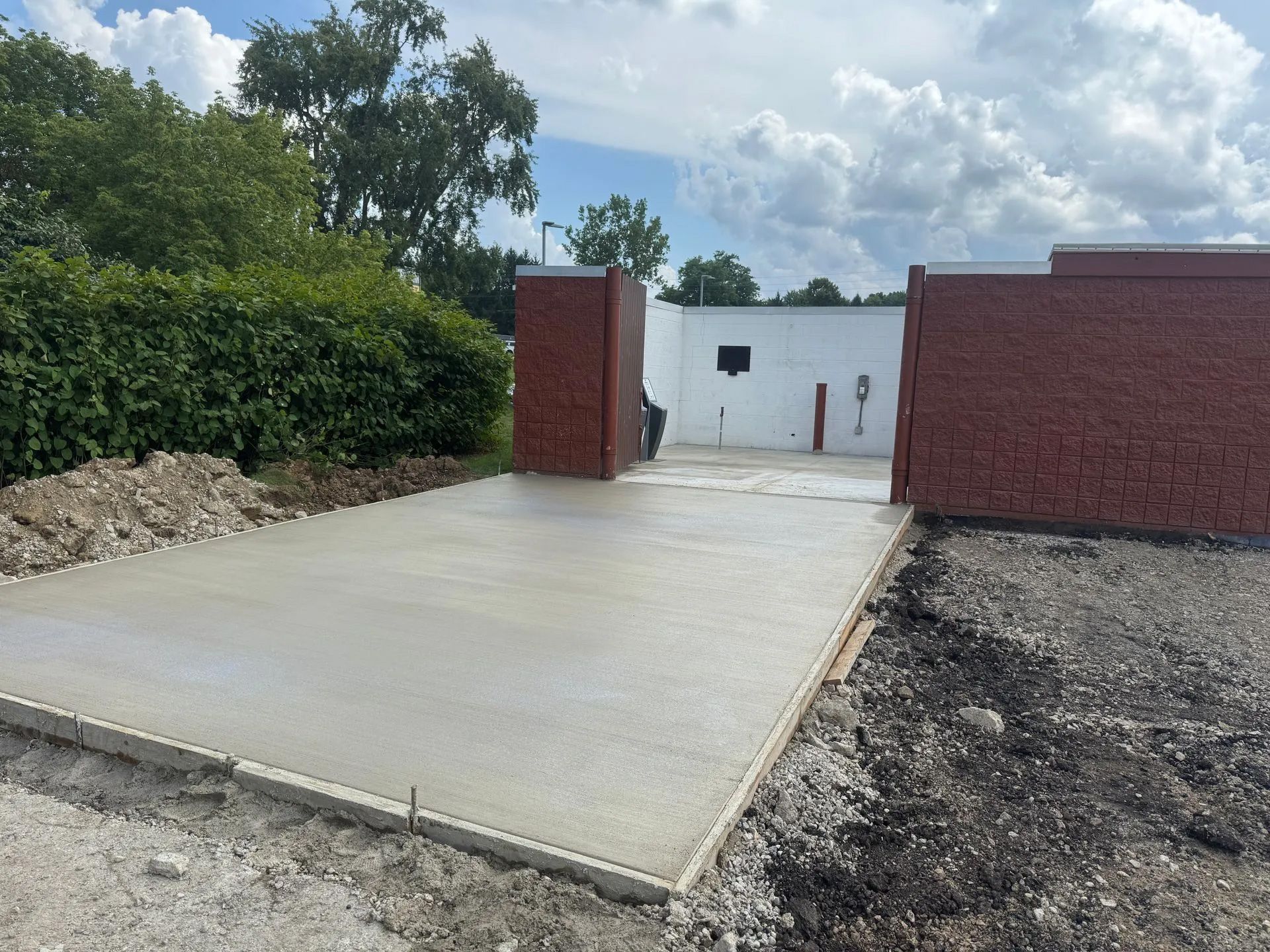 Newly poured concrete driveway leading to a white wall between two red brick pillars under a cloudy sky.