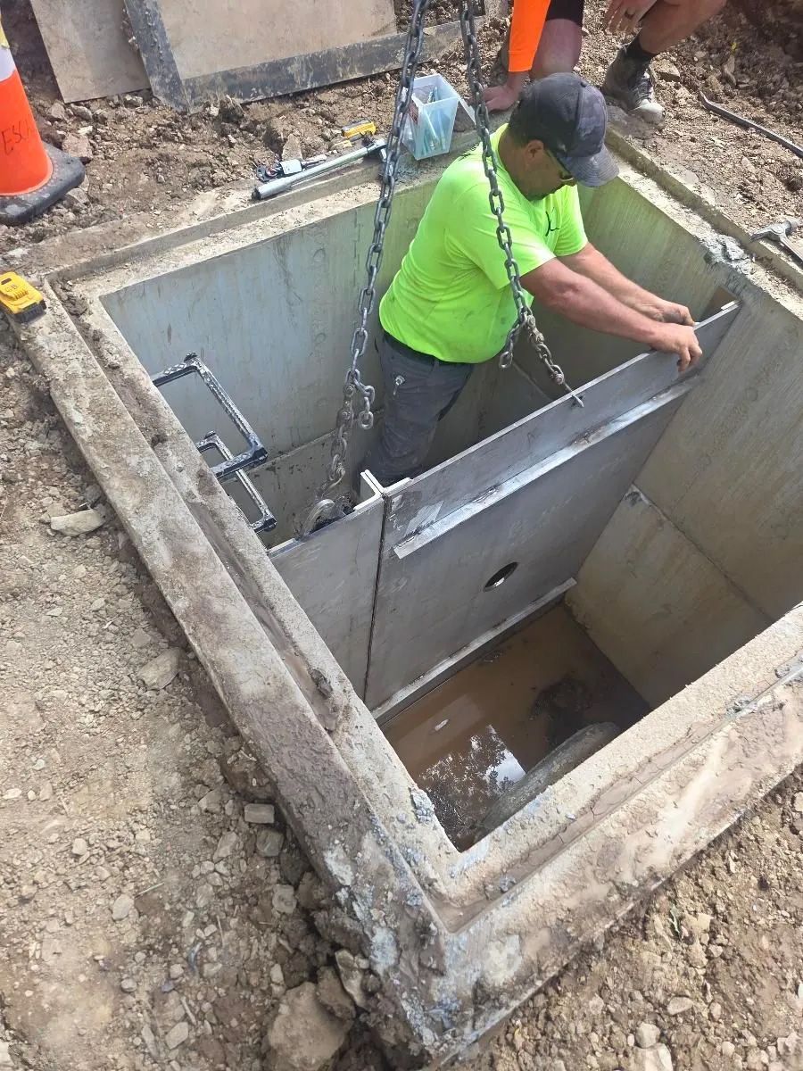 A construction worker in a neon green shirt lowers a large metal panel into a square concrete utility vault.