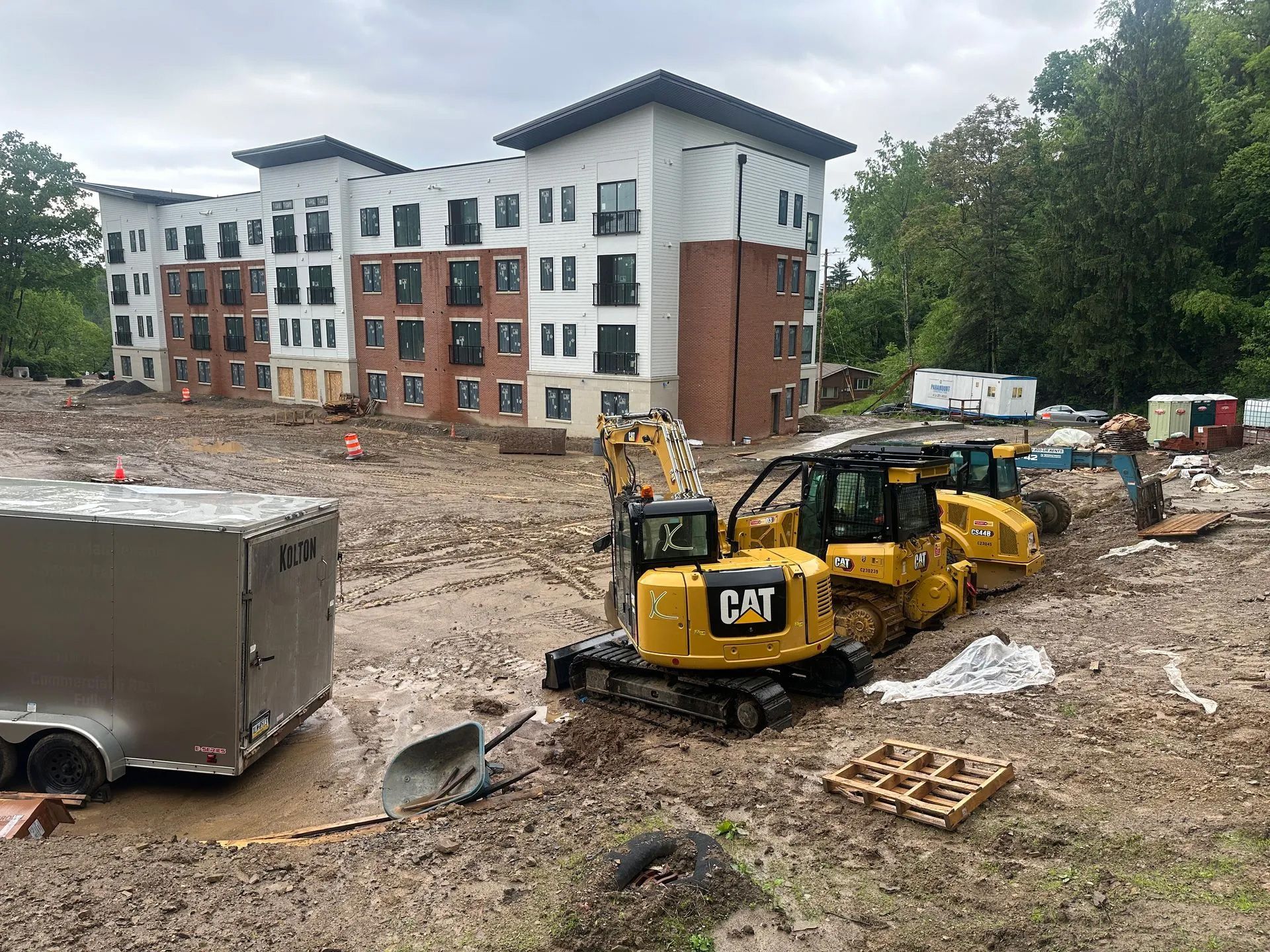 Construction site with yellow CAT excavator, dump truck, and a multi-story building in the background.