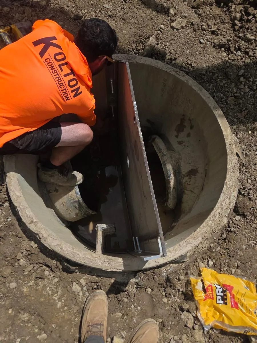 Worker in orange vest inspects a round concrete manhole with tools in a dirt excavation area