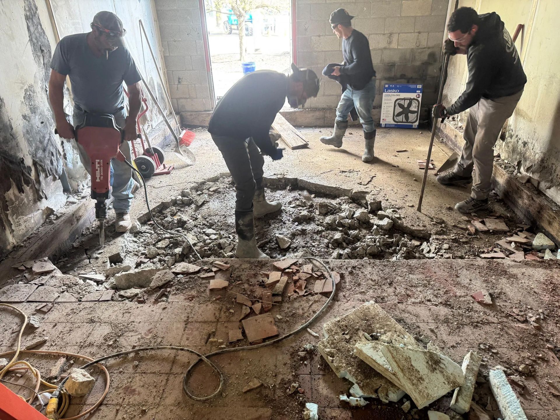 Four construction workers in an interior workspace use jackhammers and tools to break up a concrete floor.